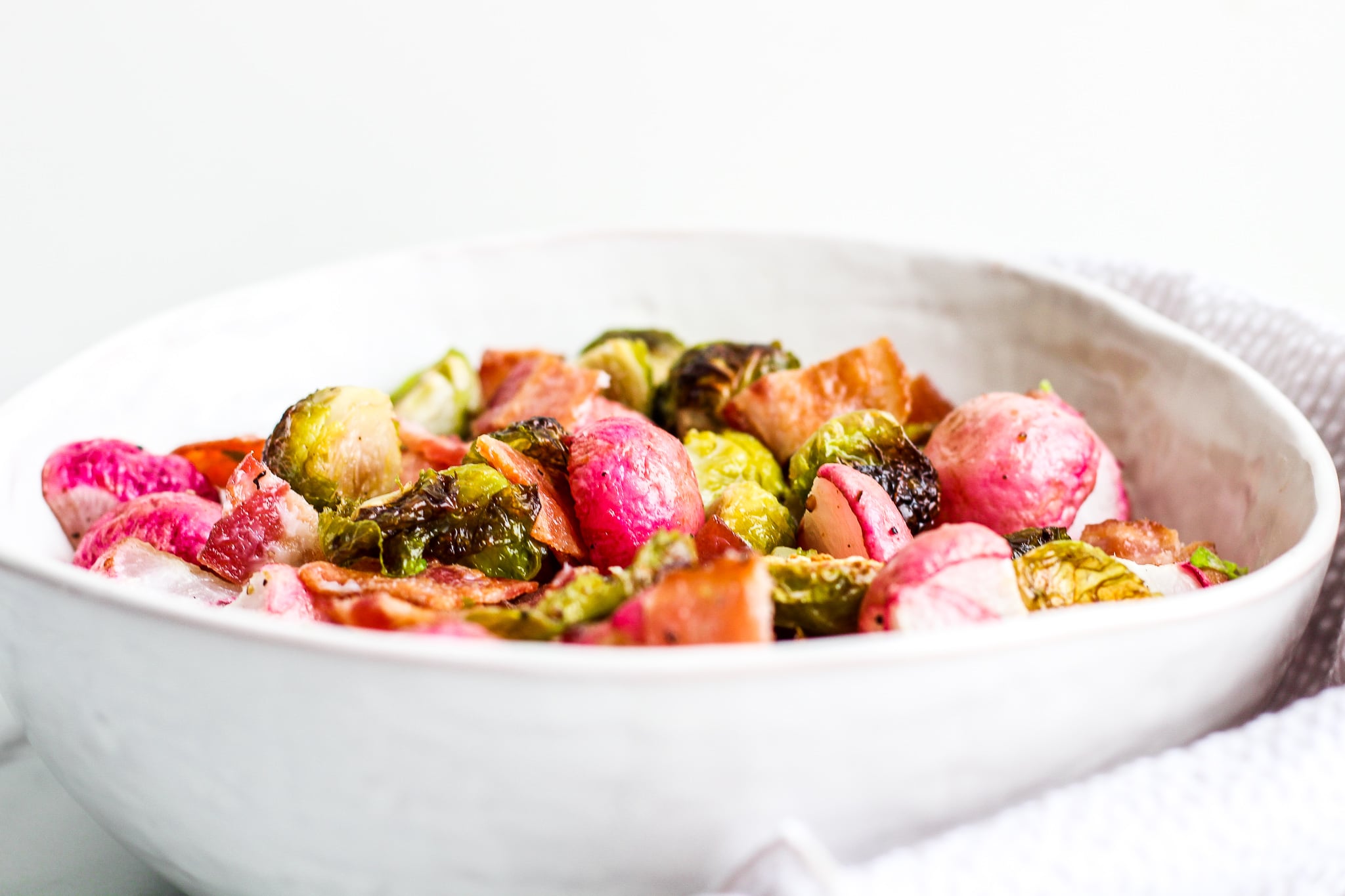 Side view of white bowl filled with roasted radishes, roasted Brussels sprouts and bacon 