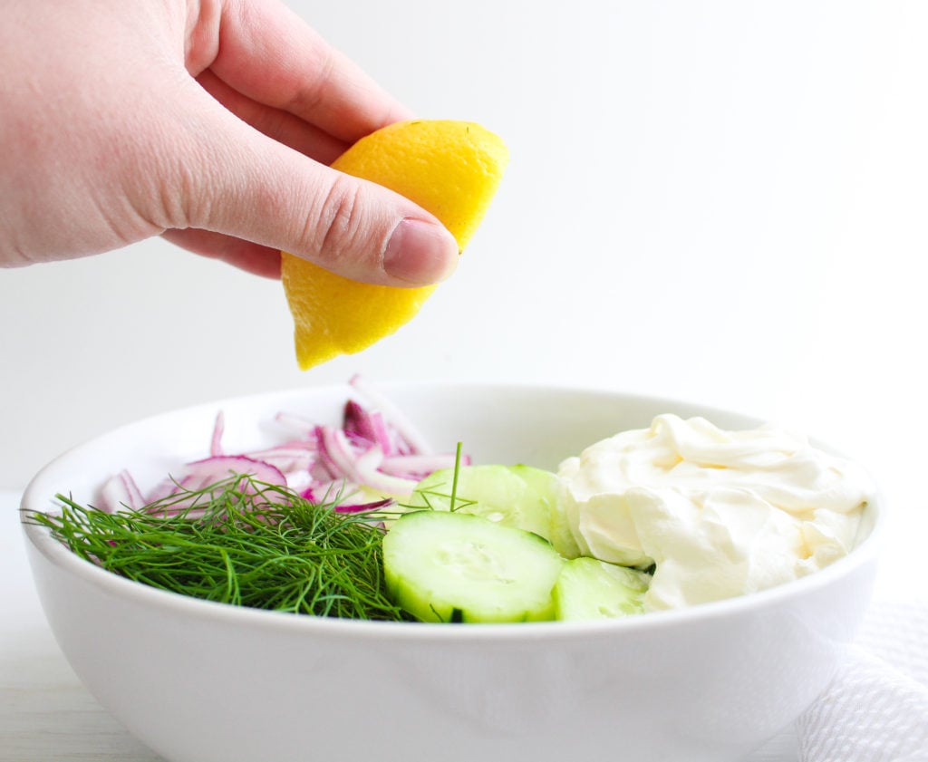 A hand squeezing lemon over a bowl with sliced cucumbers, fresh dill, red onion and sour cream 