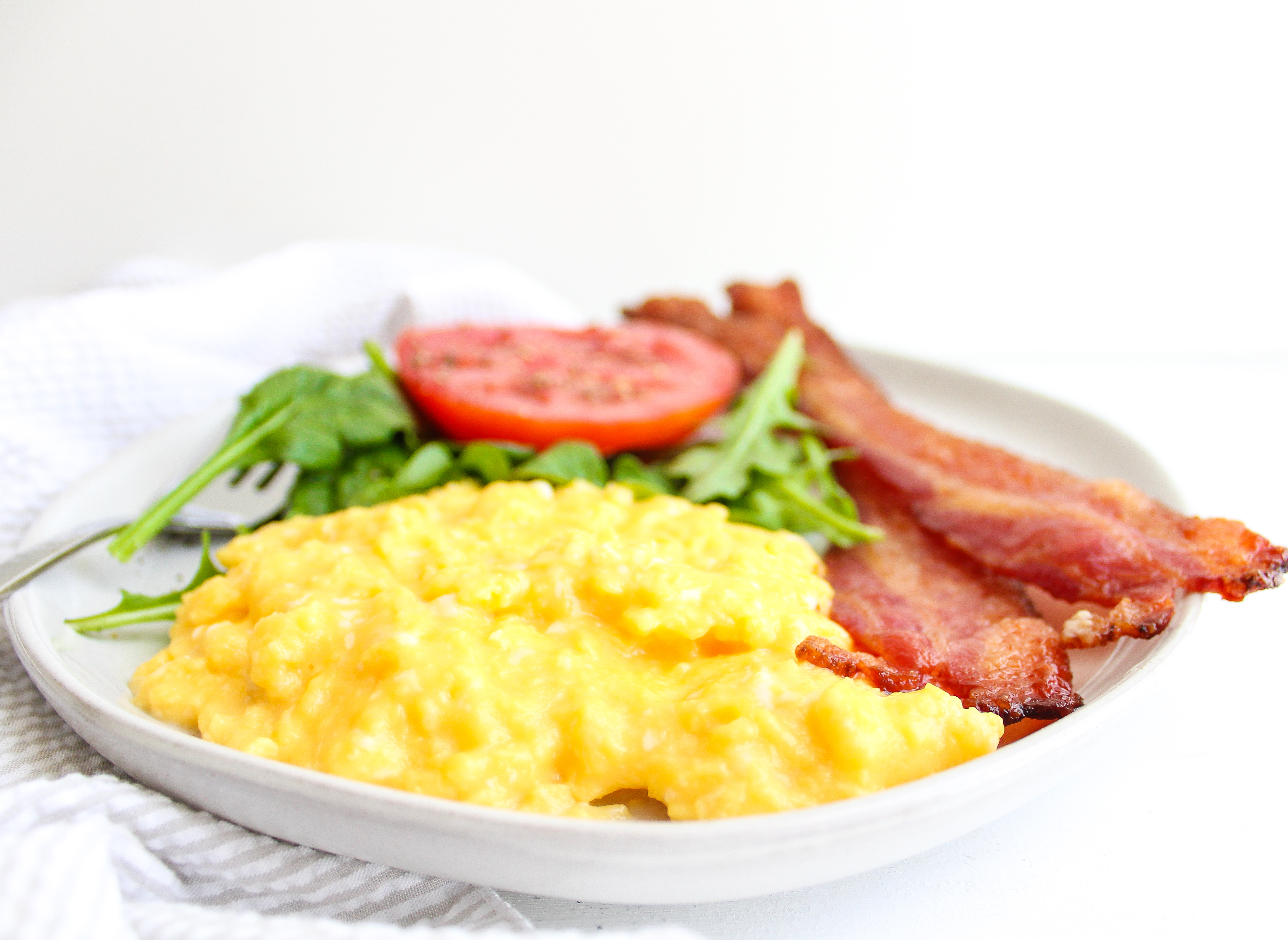 A plate filled with creamy scrambled eggs, crispy bacon, fresh greens and a slice of tomato