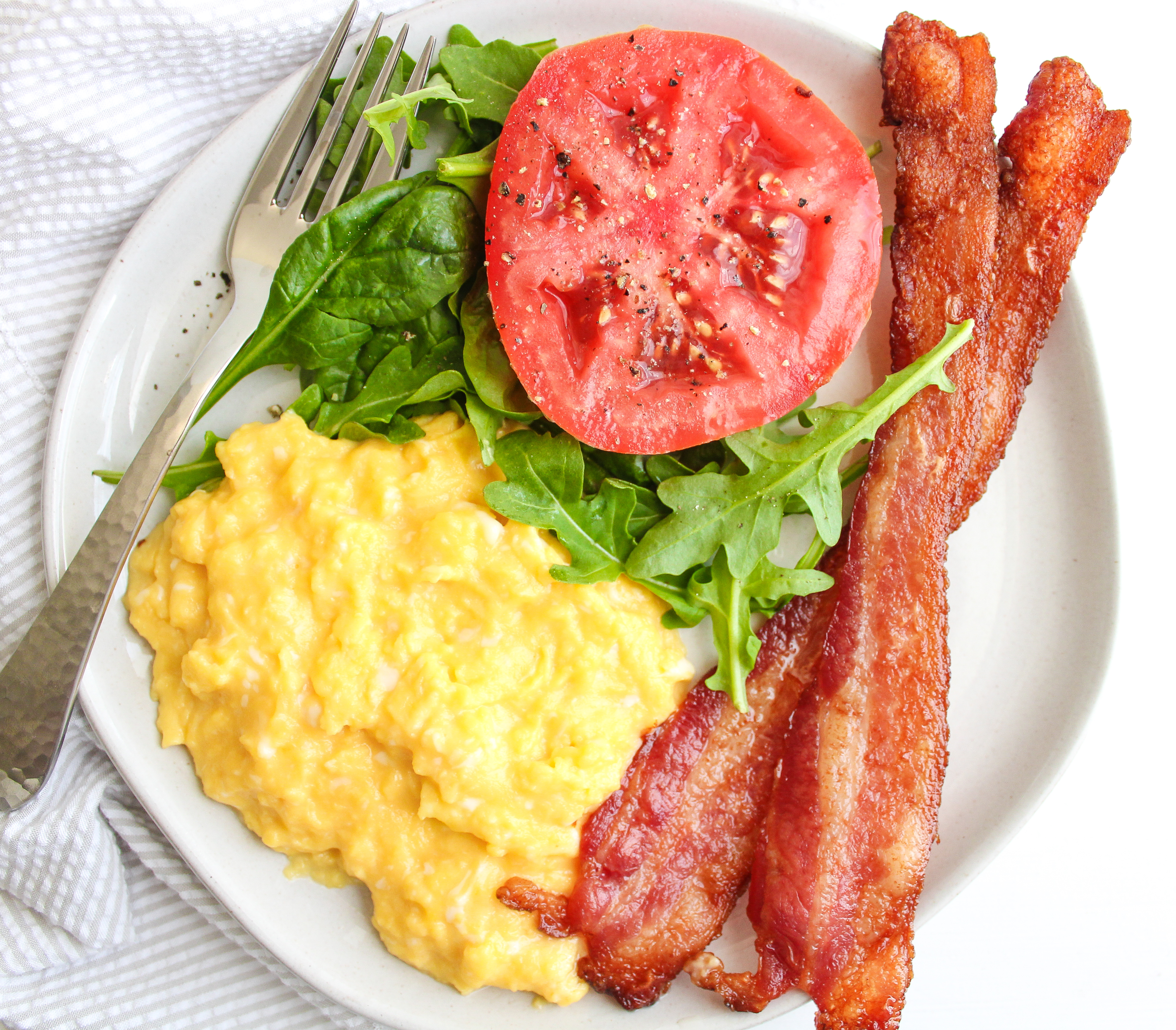 A plate filled with creamy scrambled eggs, crispy bacon, fresh greens and a slice of tomato