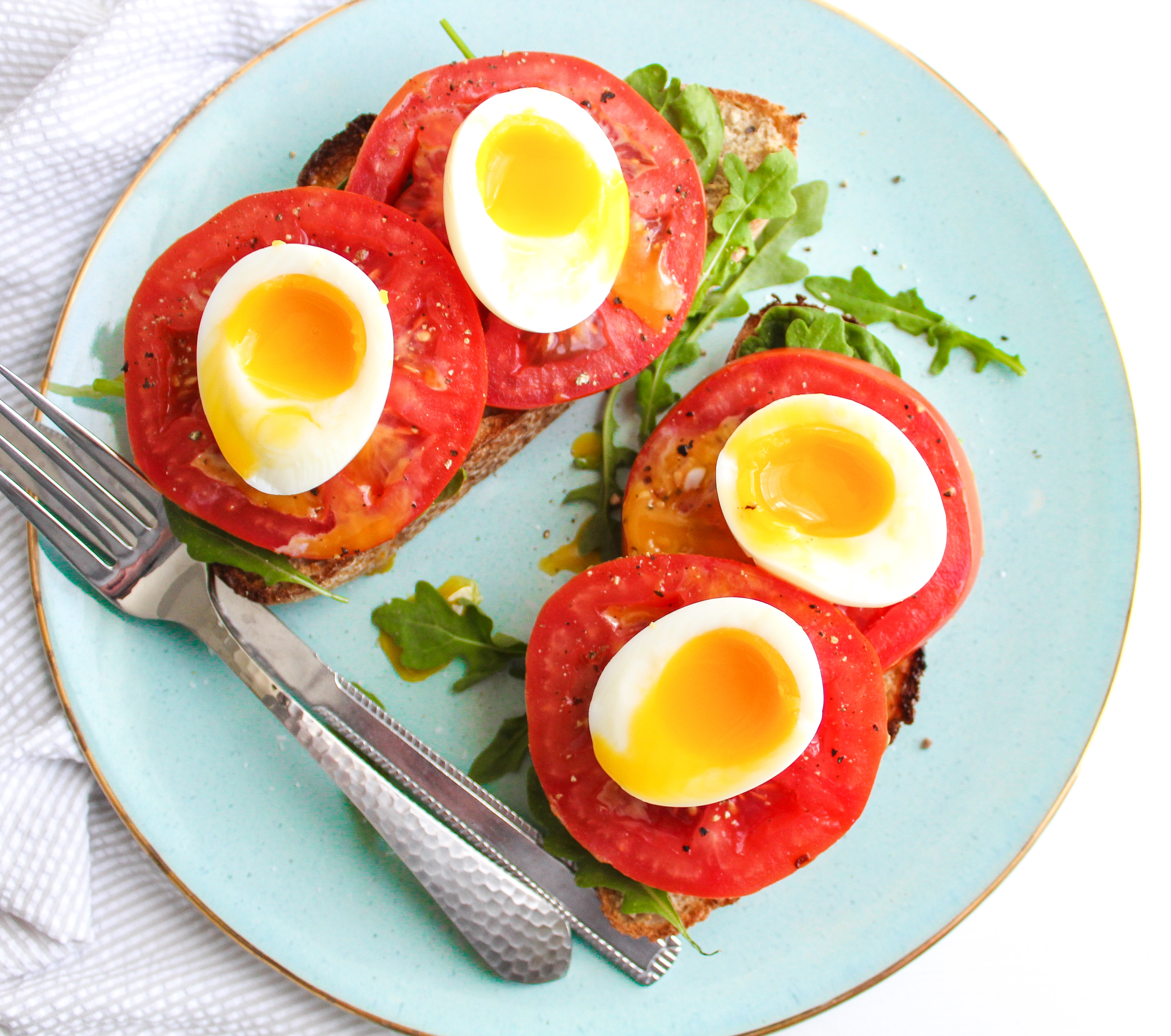 Sourdough toast topped with fresh greens, tomato slices and jammy eggs with the yolks running down