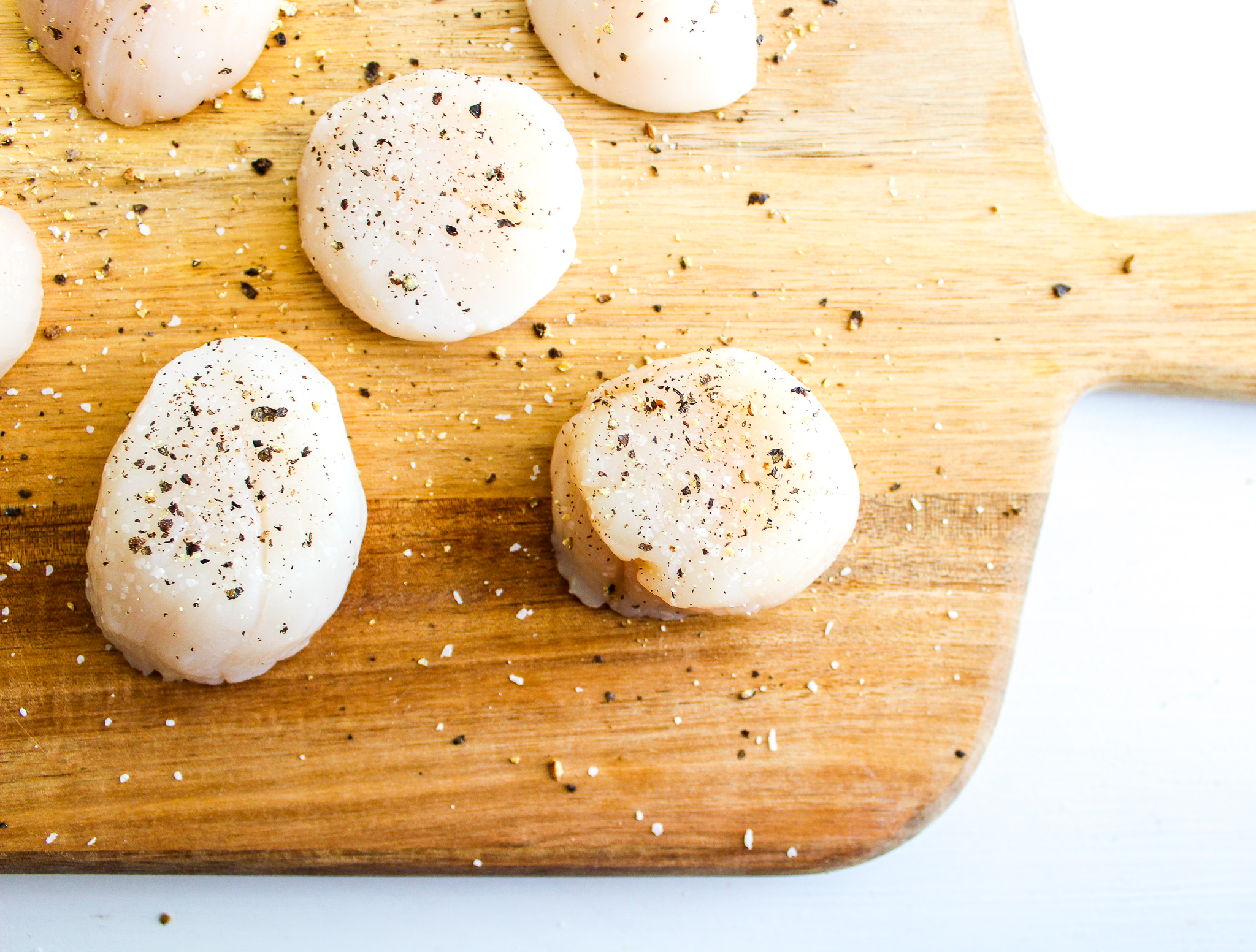 Raw scallops on a wooden cutting board being seasoned with salt and fresh cracked black pepper
