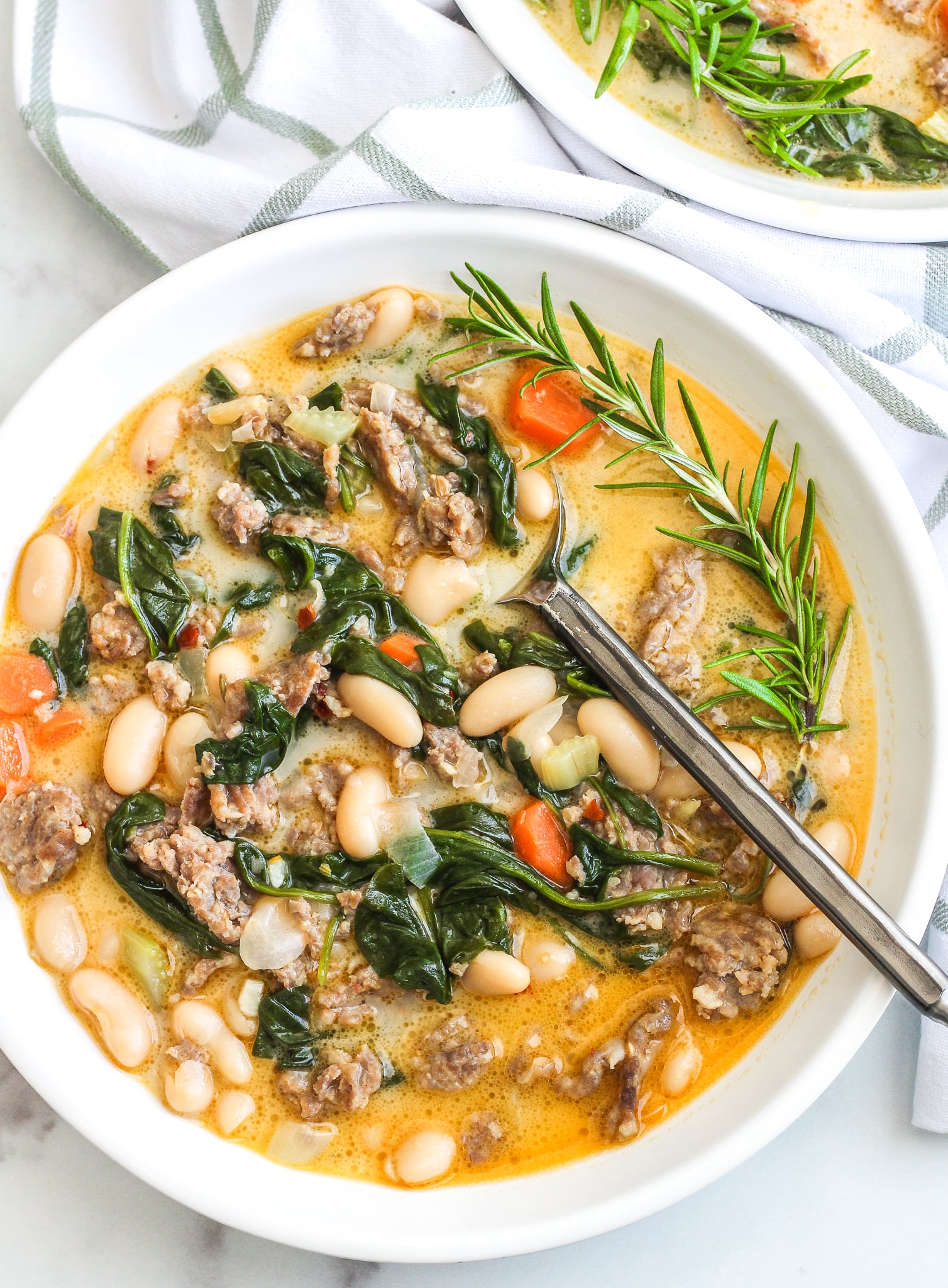 Italian White Bean Soup in a white bowl with a metal spoon and rosemary on the side 