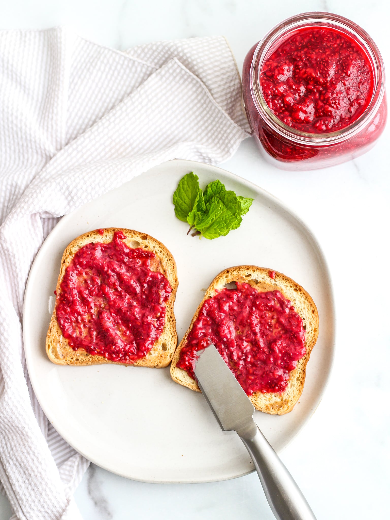 Easy Raspberry Chia Seed Jam spread on gluten free toast with a sprig of mint on the plate and a jar of the jam behind it 