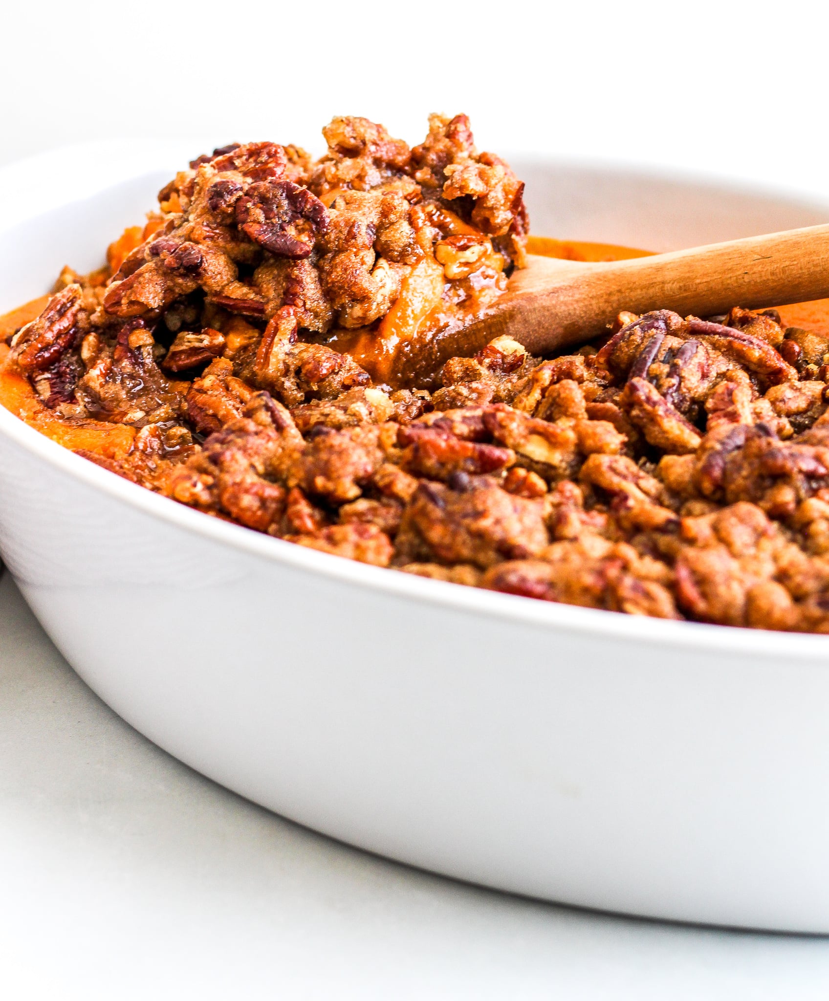 Sweet Potato Casserole topped with a pecan topping, a spoon is scooping some out of a white dish 