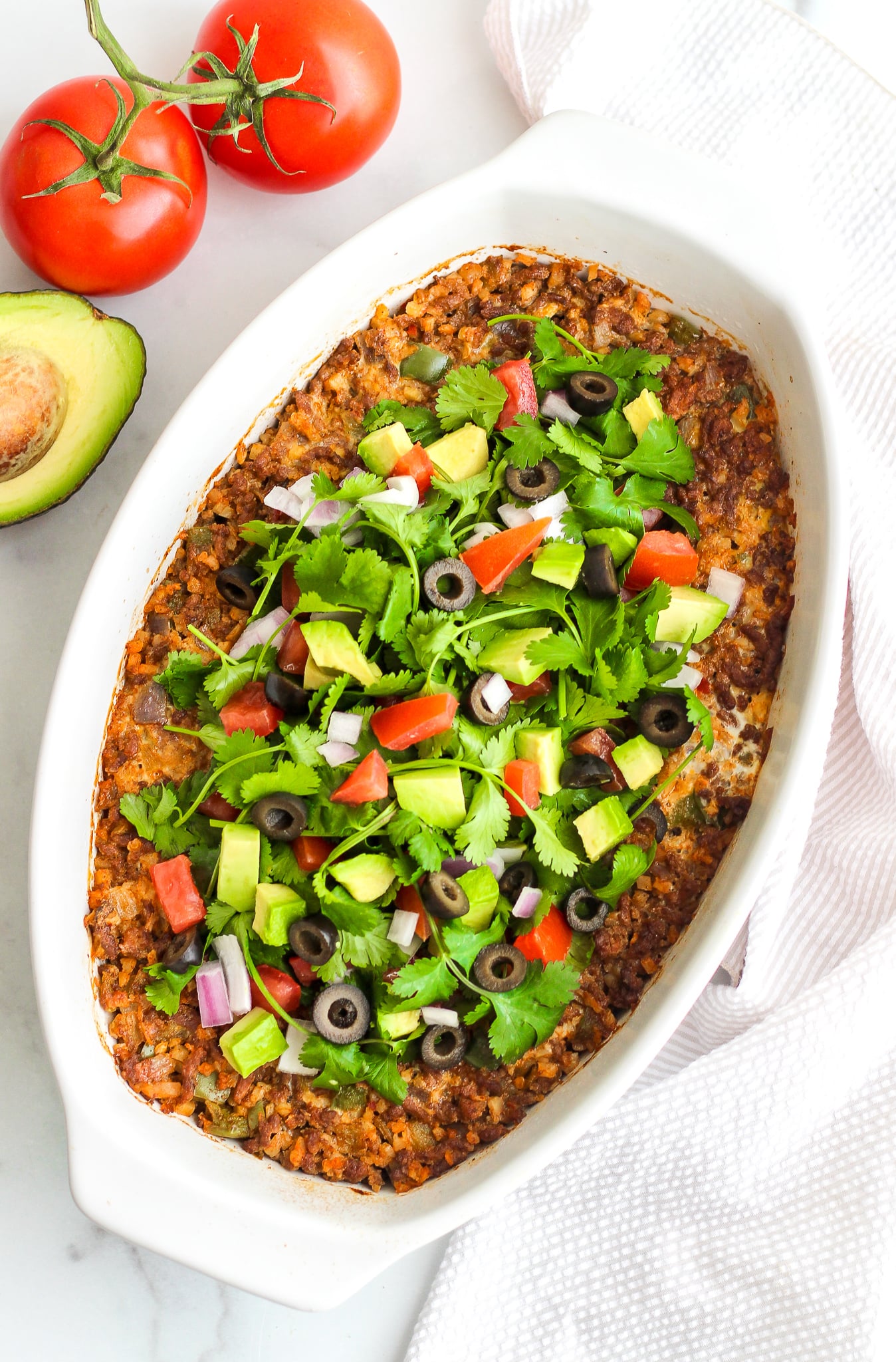 Whole30 taco bake in a white oval casserole dish topped with fresh cilantro, avocado, tomato, black olives and red onion