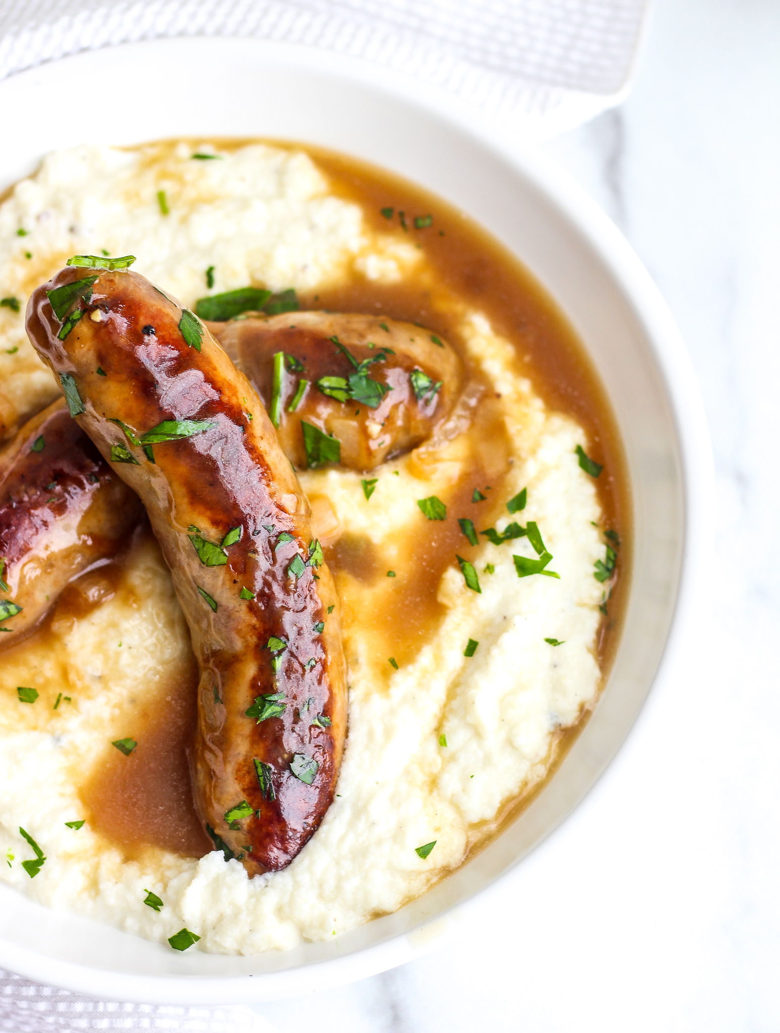 White bowl with creamy cauliflower mash topped with two browned sausages and onion gravy 
