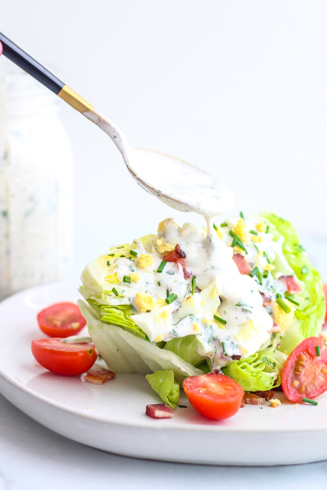A spoon drizzling homemade ranch over a wedge of iceberg lettuce topped with bacon & tomatoes 