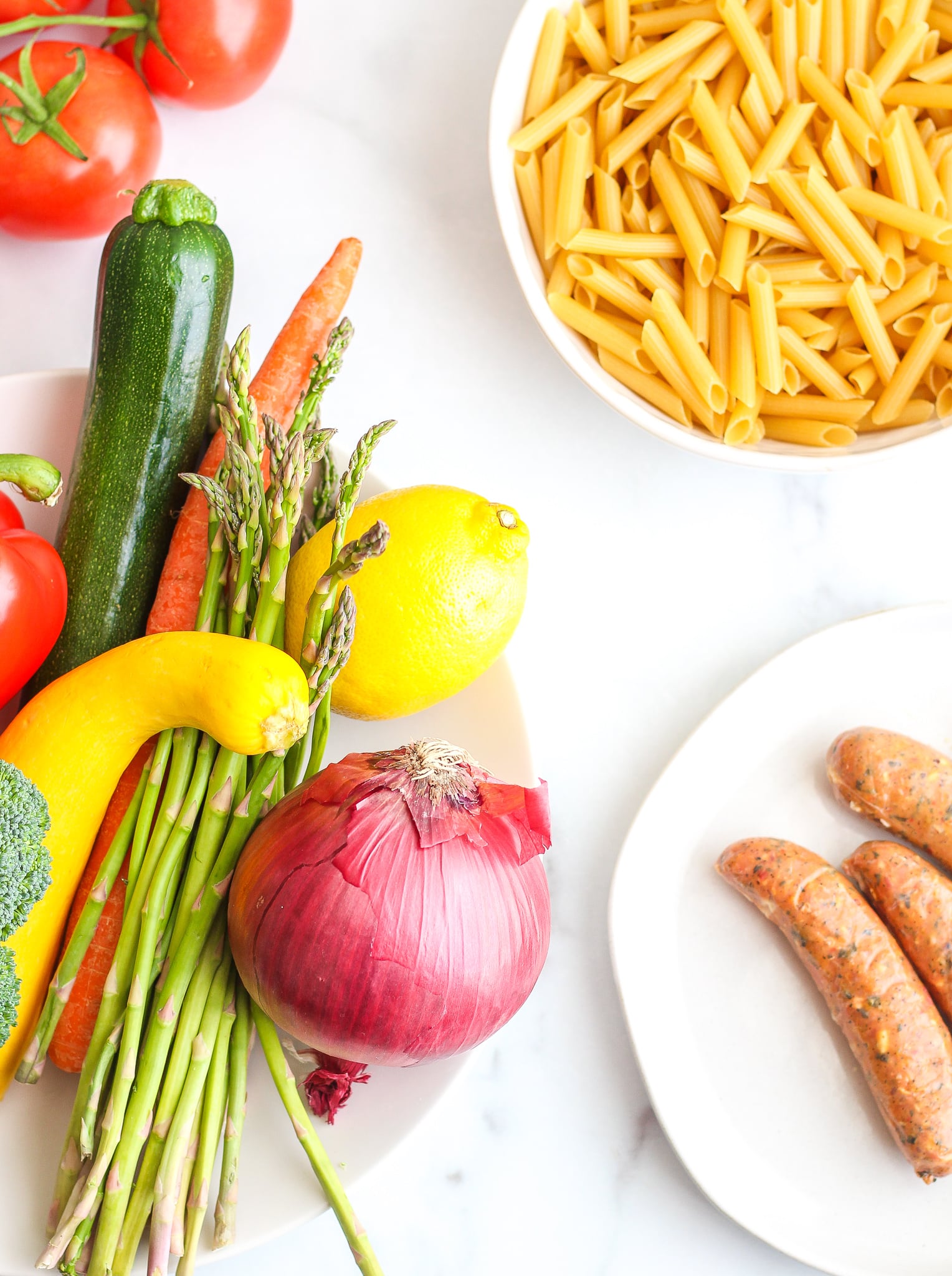 Flat lay of bowl of penne pasta, plate filled with veggies, tomatoes on the vine and chicken sausages 