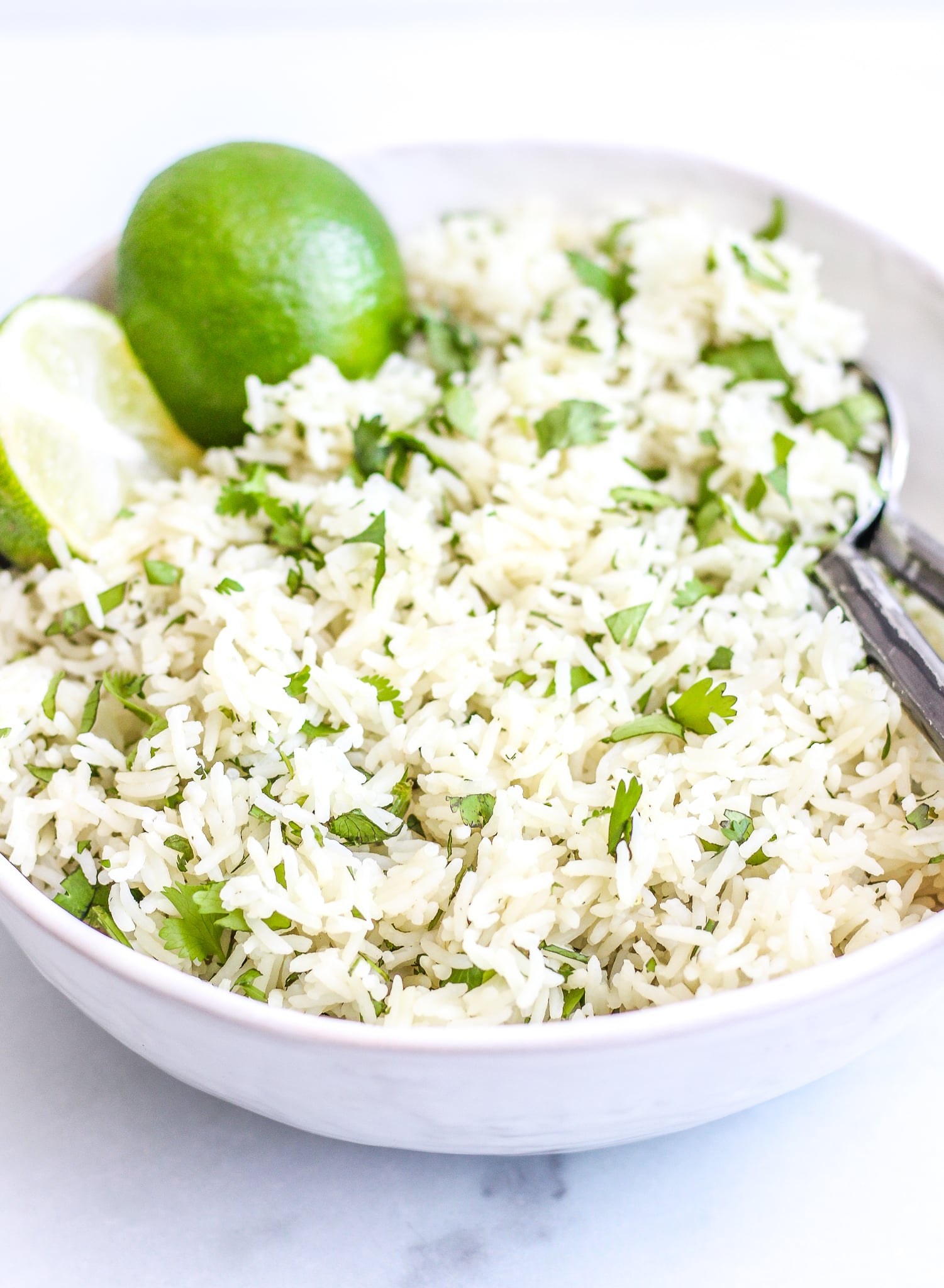 Side view of fluffy cilantro lime rice in a white serving bowl with halved limes as a topping and serving spoons on the side of the bowl 