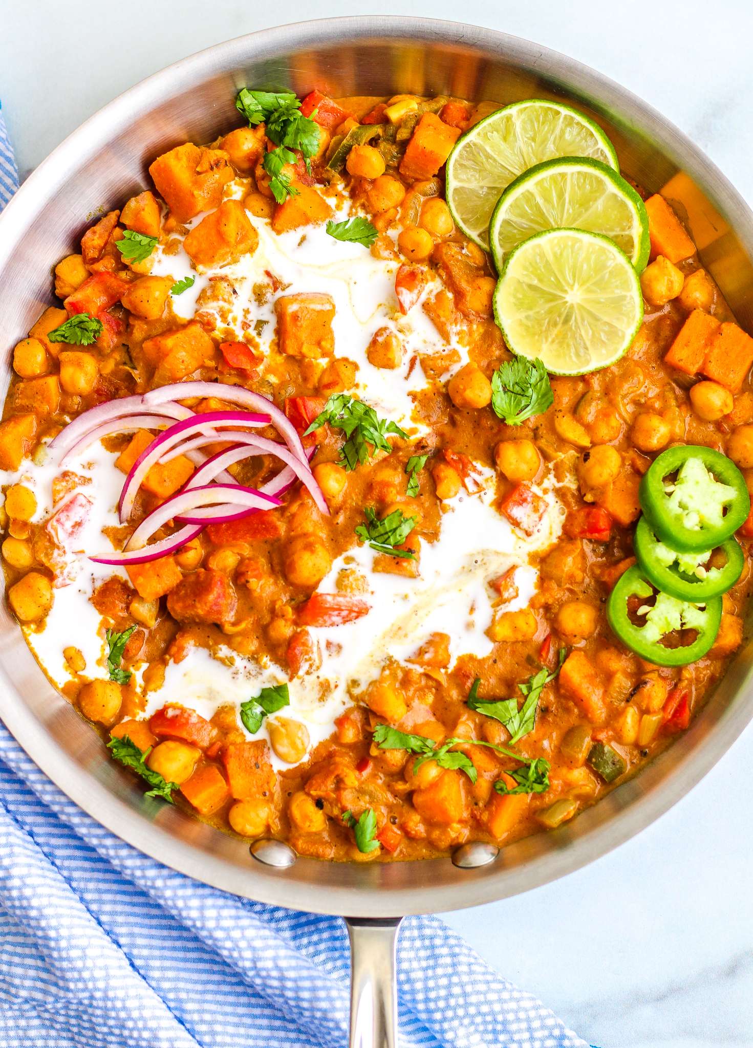 Chickpea curry in a skillet. The curry has swirls of coconut milk in it and is topped with thinly sliced limes, jalapeños, and red onion. It is also sprinkled with fresh chopped cilantro. 
