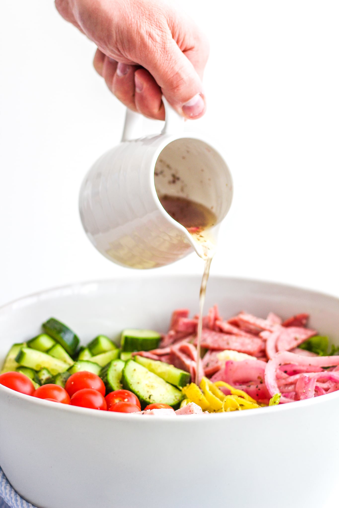 Italian chopped salad in a large ceramic bowl with layers of cherry tomatoes, cubed ham, chopped salami, chopped cucumbers, onions and lettuce with hand pouring Italian dressing over it 