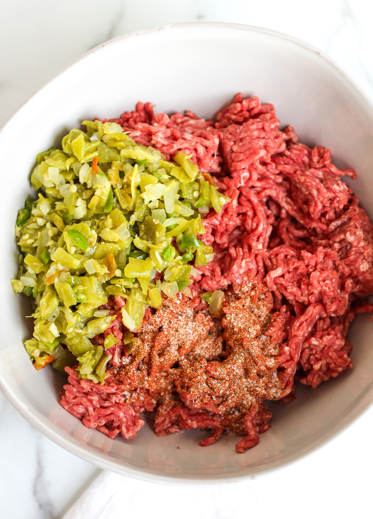 Large ceramic mixing bowl filled with ground beef, a mixture of green chiles, onions, garlic and jalapeño along with a blend of spices 