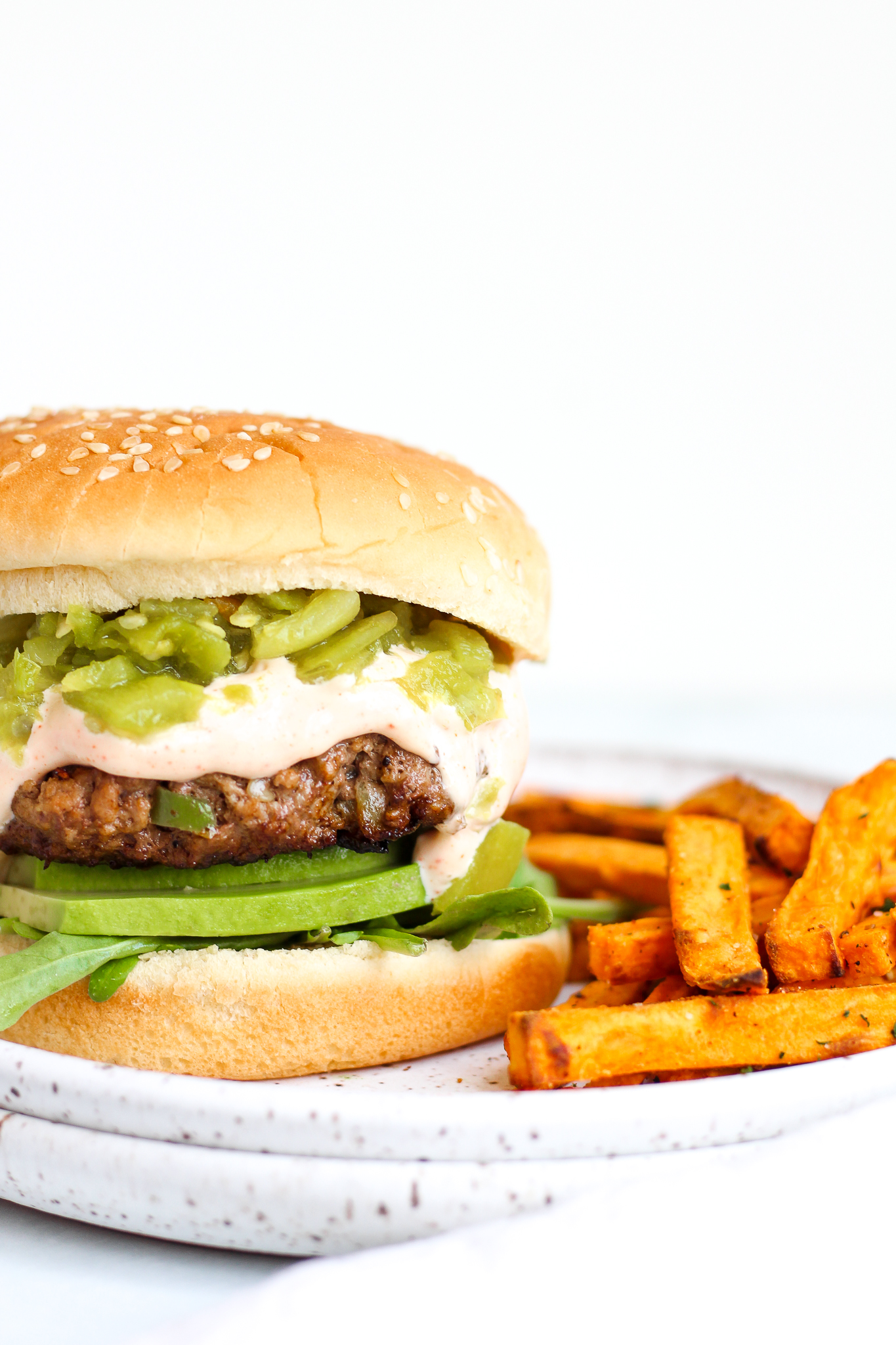 Green Chile burger on a sesame seed bun on a white ceramic plate served alongside homemade sweet potato fries 