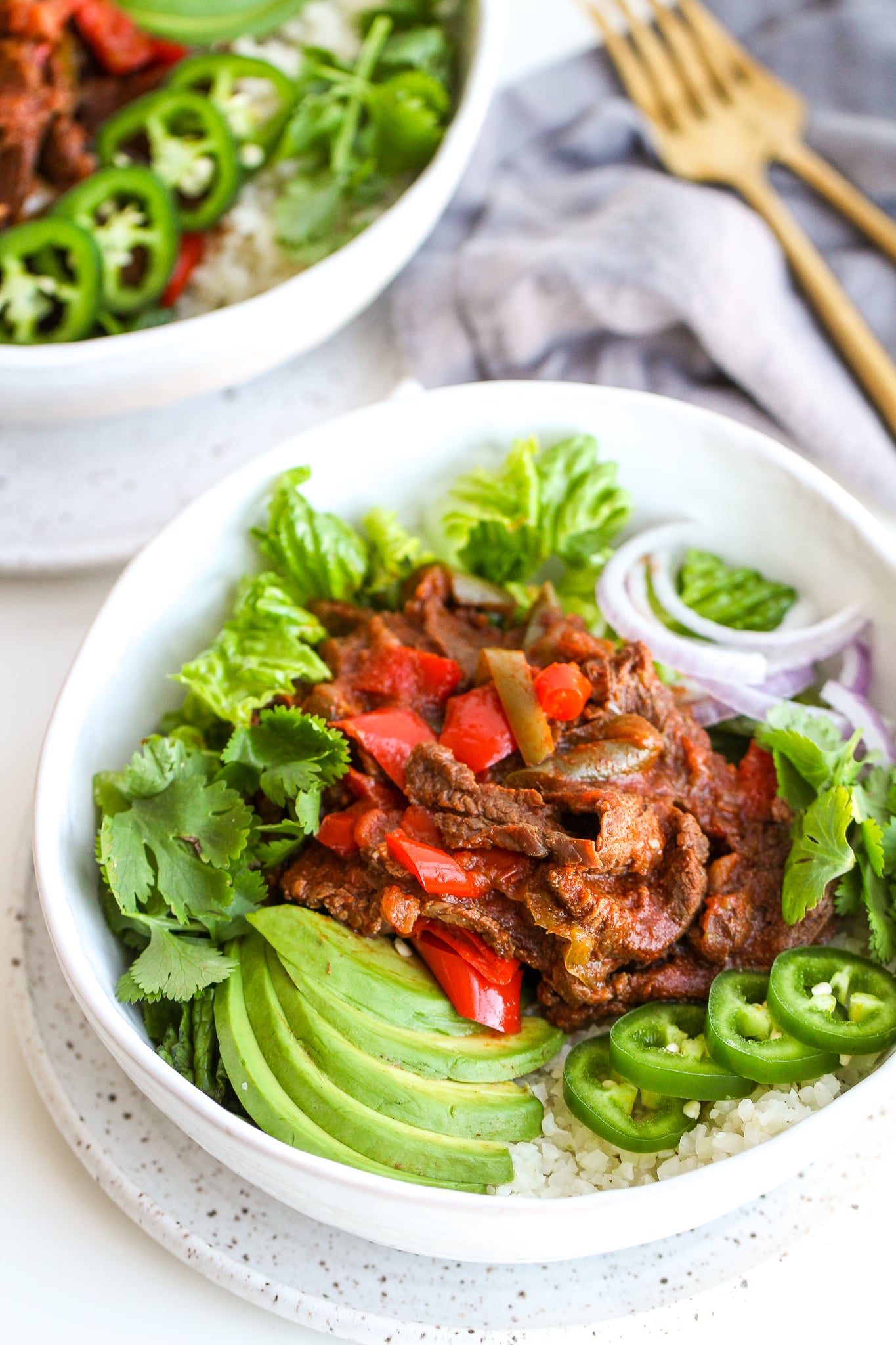 Instant Pot Fajita Steak and Peppers in a bowl with lettuce, cauliflower rice, sliced avocado, jalapeño and fresh cilantro in a white bowl 
