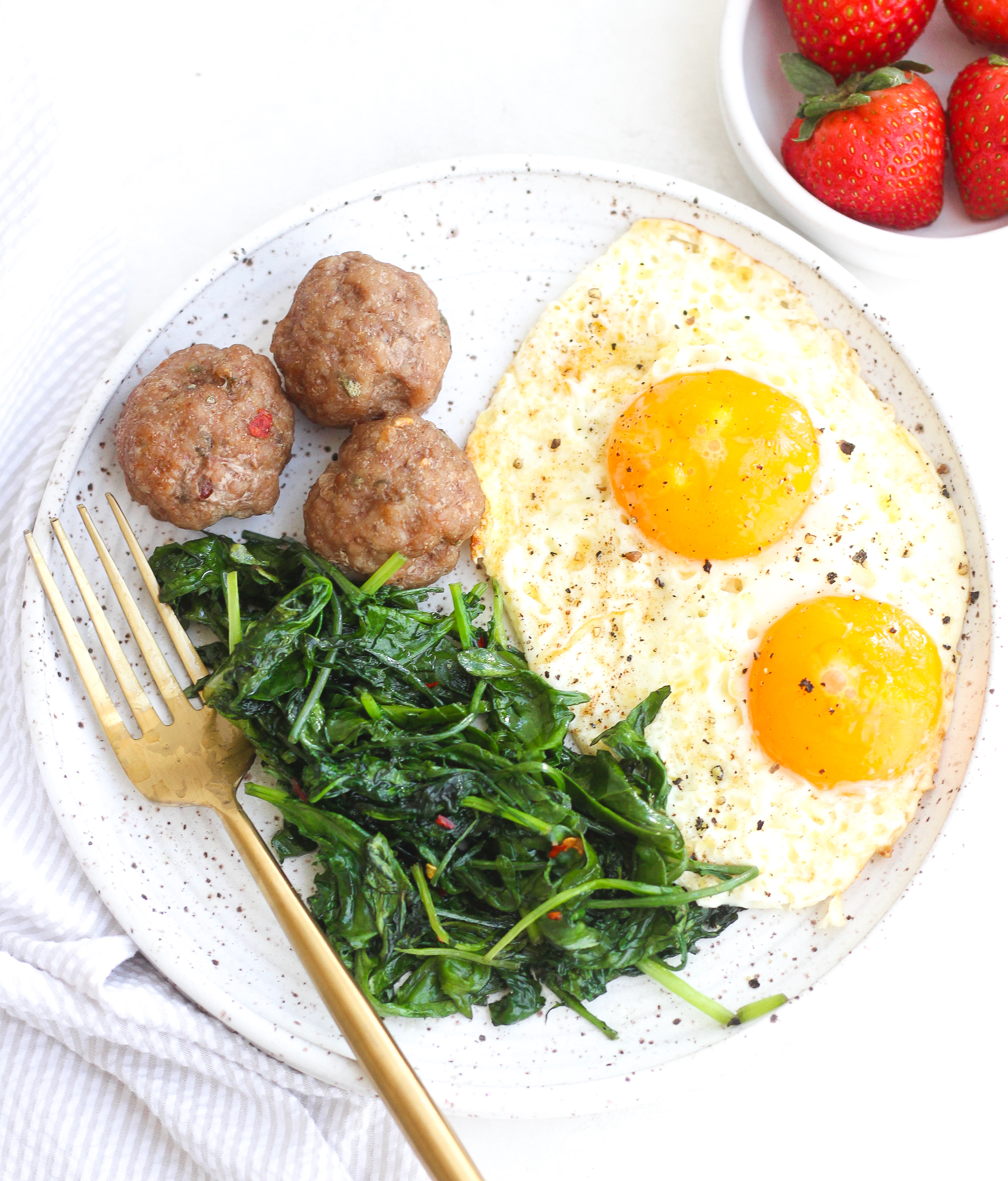 breakfast meatballs on a ceramic plate served alongside sautéed kale and sunny side up eggs