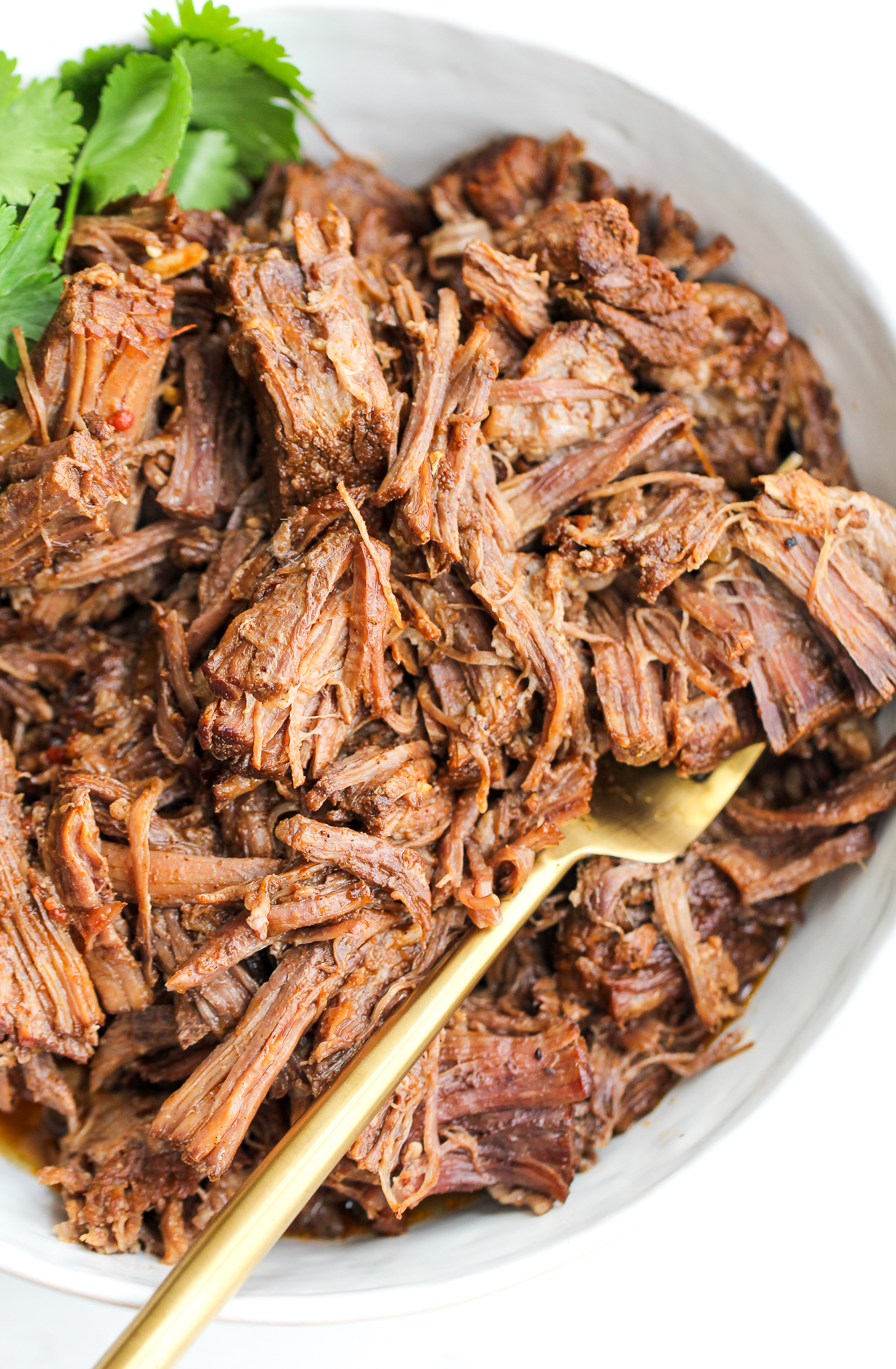Instant Pot Mexican shredded beef in a white serving bowl with a gold fork and fresh cilantro on top 