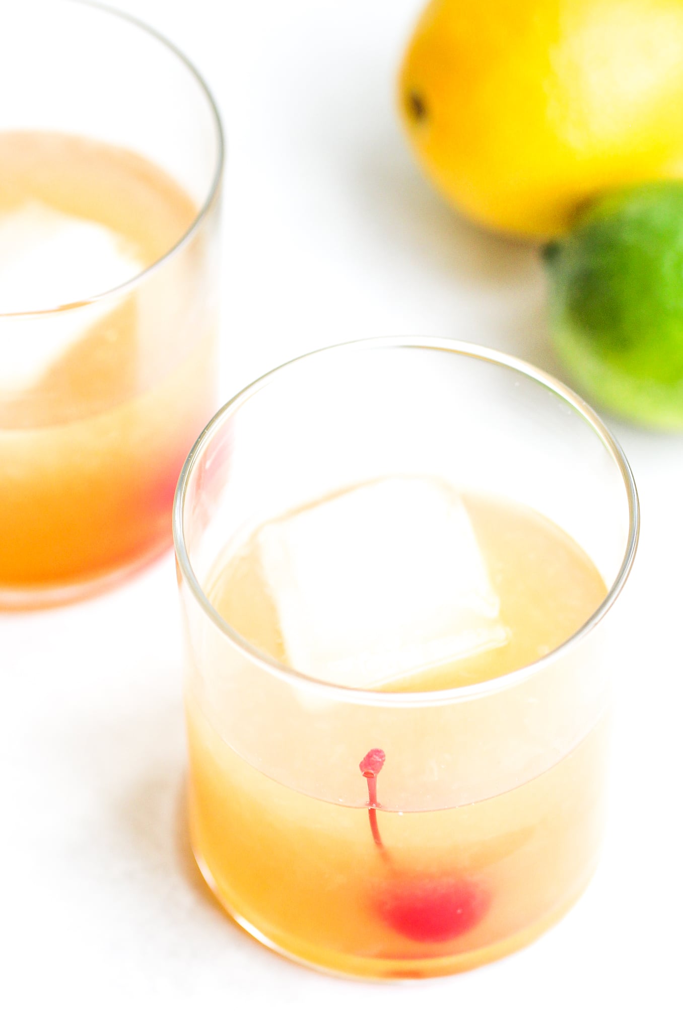 overhead shot of a classic whiskey sour in a clear cocktail glass. You can see a large ice cube and maraschino cherry inside - lemons and limes sit in the background 