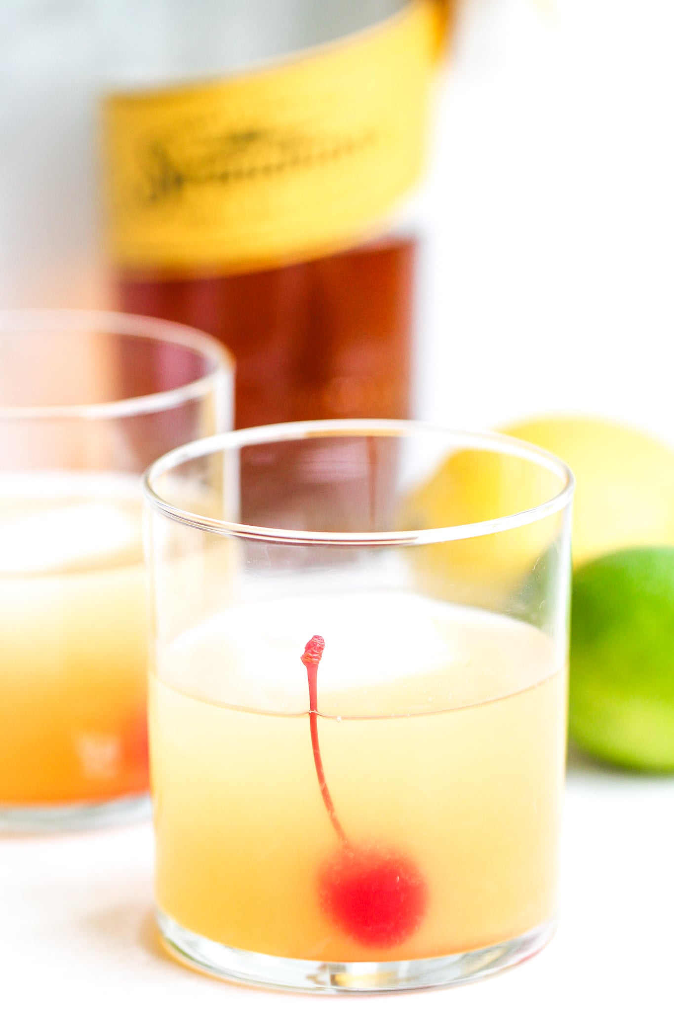 Classic whiskey sour in a clear cocktail glass with ice and a maraschino cherry. whiskey bottle stands in the background 
