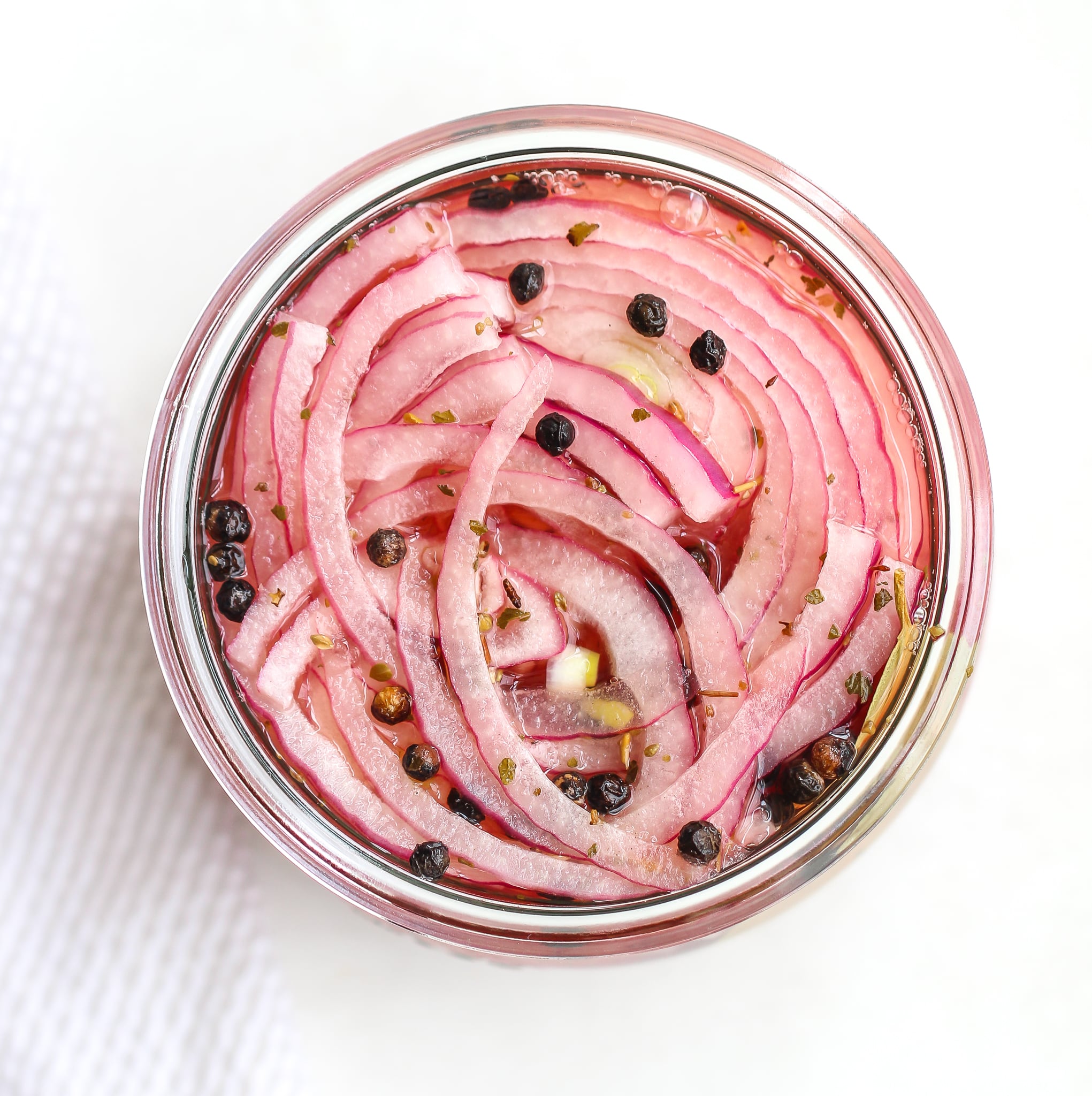 overhead shot of quick pickled red onions in a clear glass jar, there are peppercorns throughout 