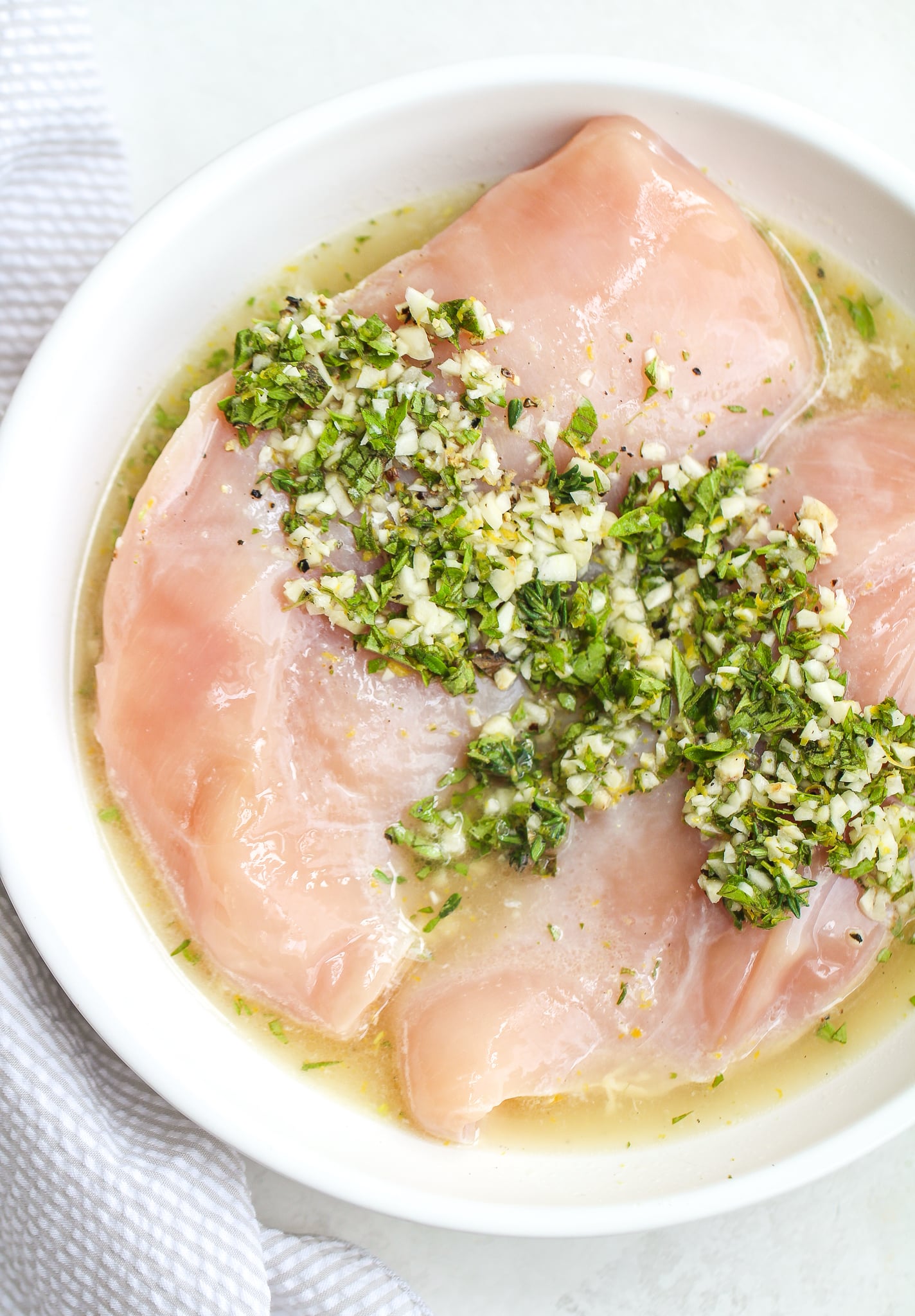 chicken breasts in a white bowl covered with greek marinade (chopped oregano and thyme with lemon zest, lemon juice, and garlic) 