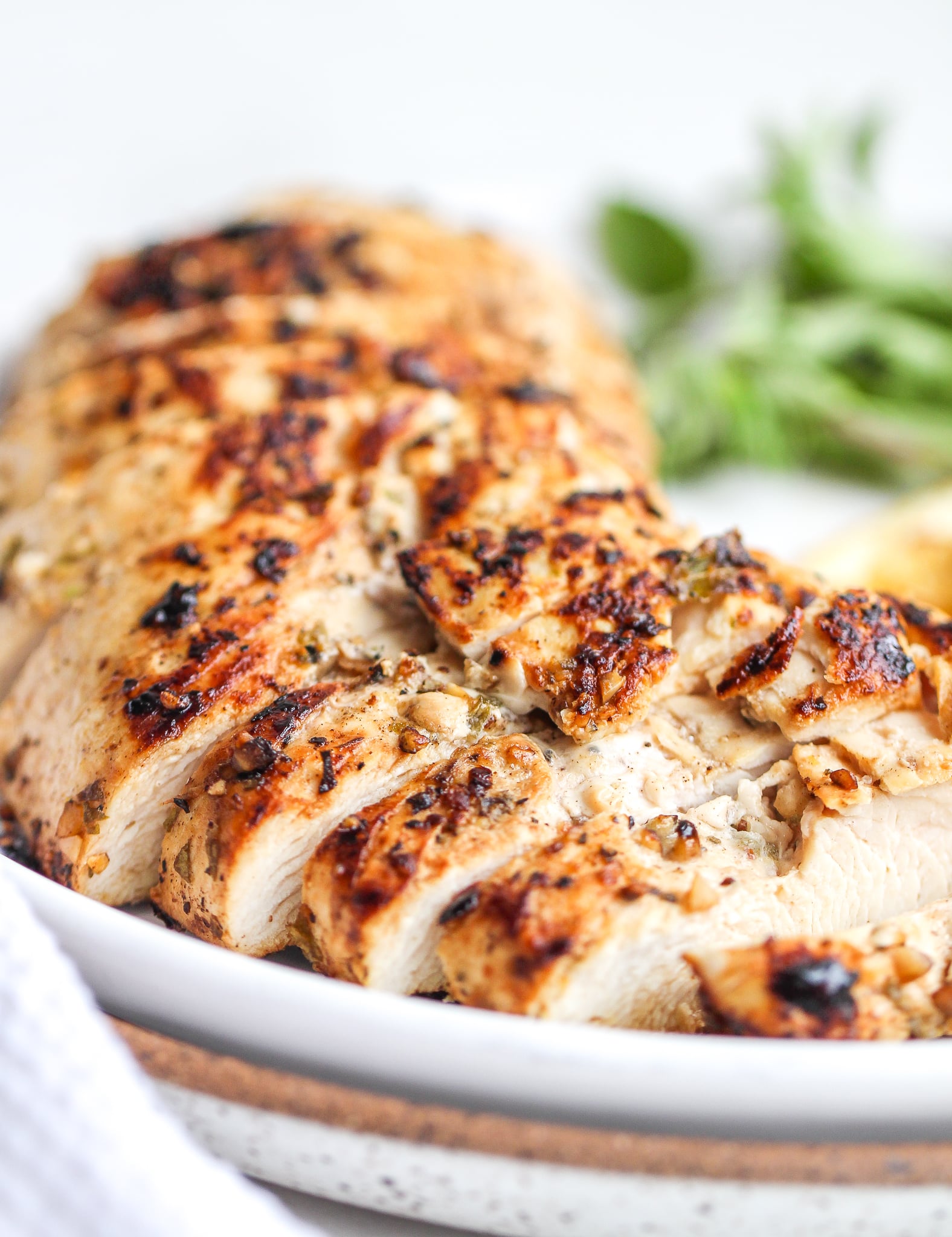close up of sliced marinated greek chicken breast on a white plate with fresh oregano in the background 
