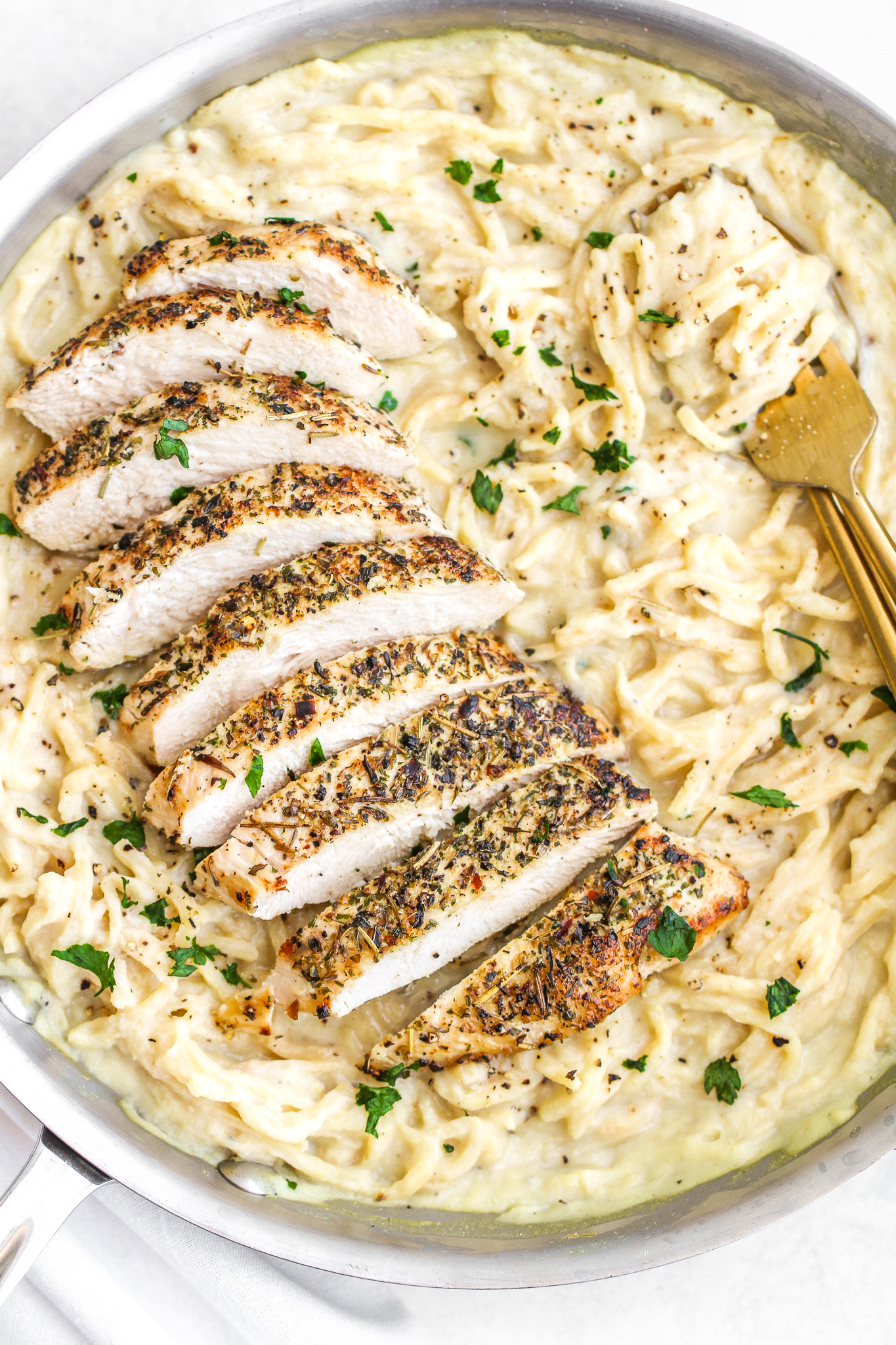 roasted garlic alfredo sauce tossed with low carb noodles in a large stainless steel pan. It is topped with Italian seasoned sliced chicken and has a gold fork swirled into it 