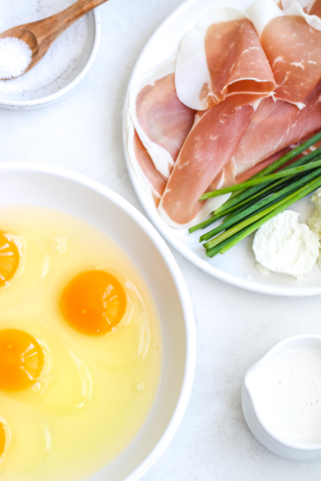flat lay with raw eggs in a bowl, a ceramic cup with cream. Prosciutto, chives and goat cheese on a white ceramic plate and a sat dish with a wooden serving spoon 