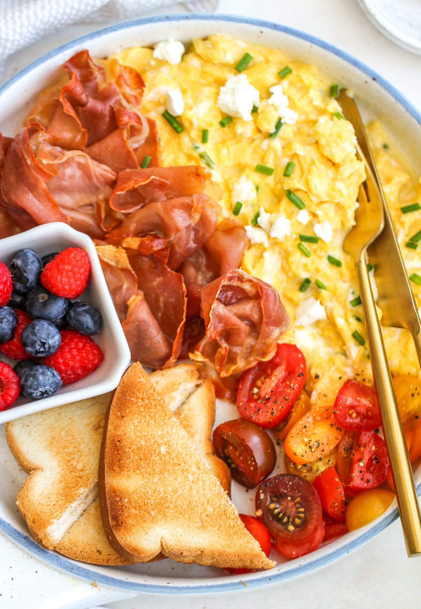 Creamy scrambled eggs on a serving platter topped with goat cheese and chives. Served alongside crispy prosciutto, a bowl of fresh berries, toast slices, and grape tomatoes sprinkled with fresh cracked pepper 