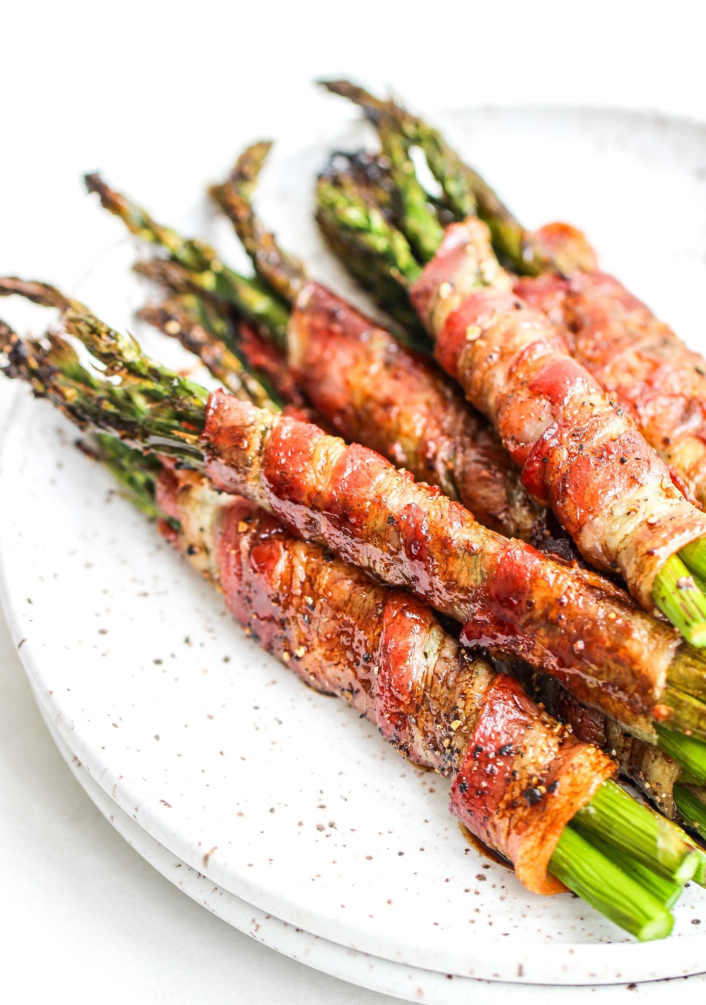 close up bacon wrapped asparagus bundles drizzled with balsamic reduction stacked on a ceramic plate with gold forks on the side of the plate 