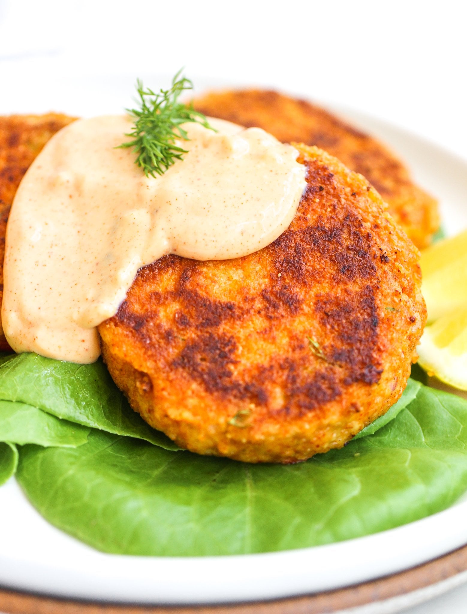 spicy garlic aioli drizzled over tuna cakes topped with fresh dill. The tuna cakes are stacked on top of a bed of butter lettuce with lemon wedges on the side 
