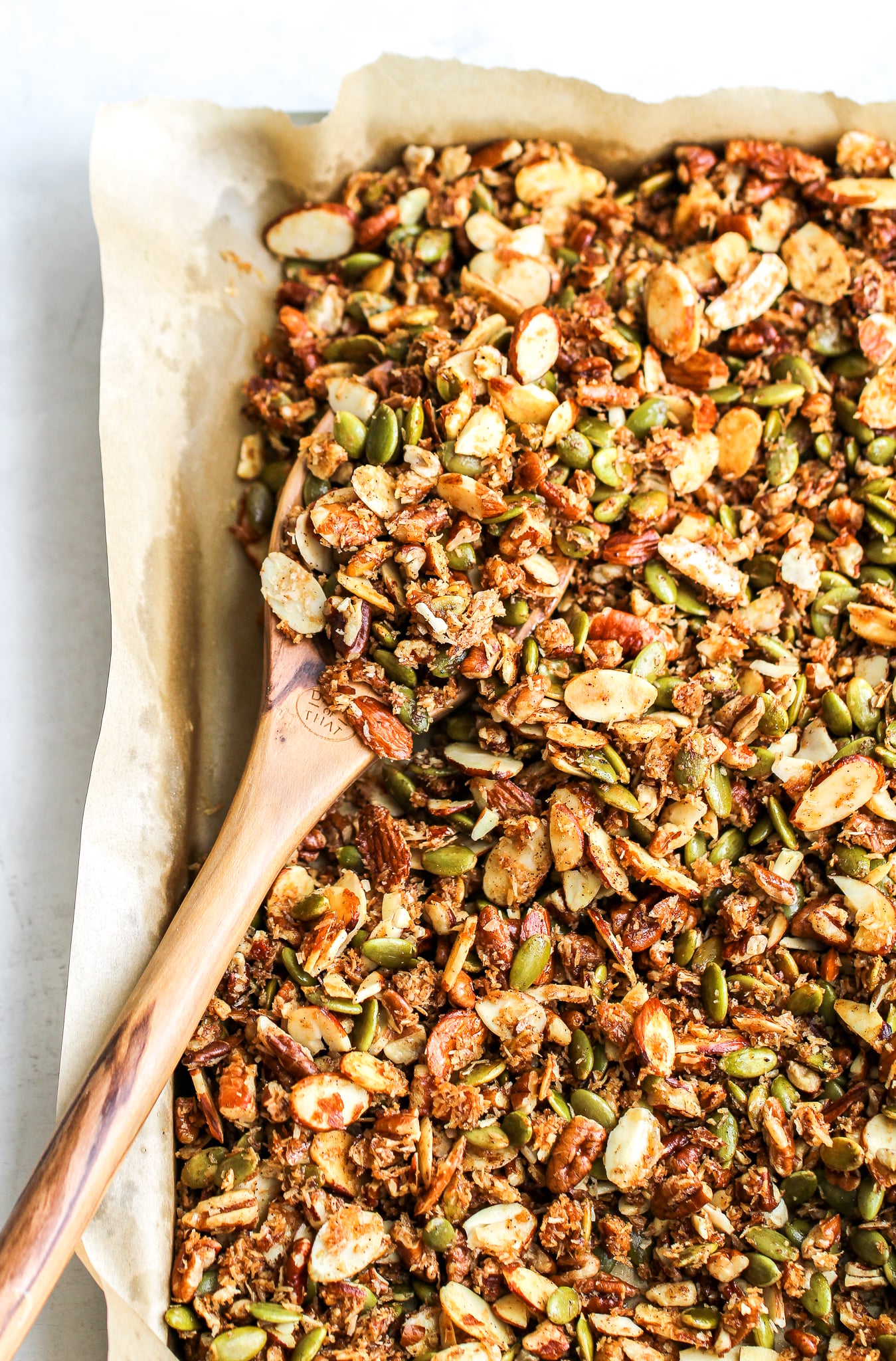 paleo granola on a parchment lined baking sheet after it has been baked. there is a wooden spoon scooping out some of the granola 