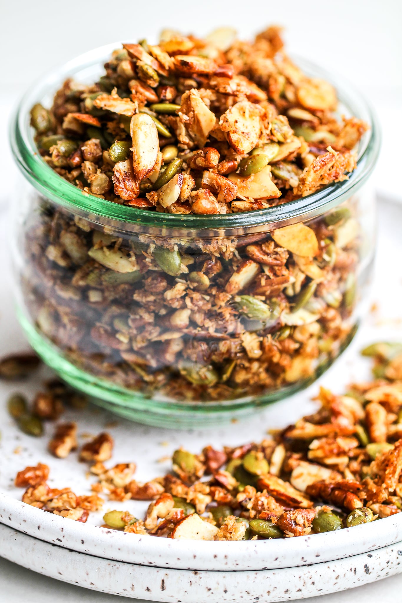 paleo granola in a glass storage jar, sitting on a speckled ceramic plate with some granola on the plate as well 
