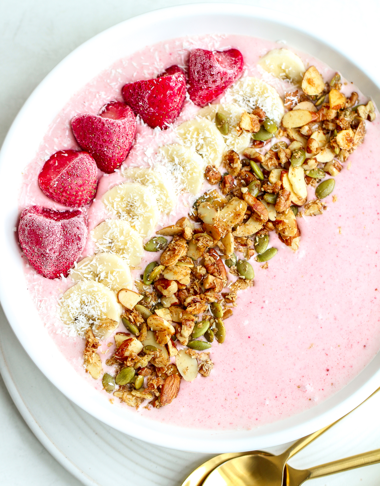 Pink strawberry banana smoothie mixture in a white bowl, topped with strawberries, banana slices, homemade granola and shredded coconut 