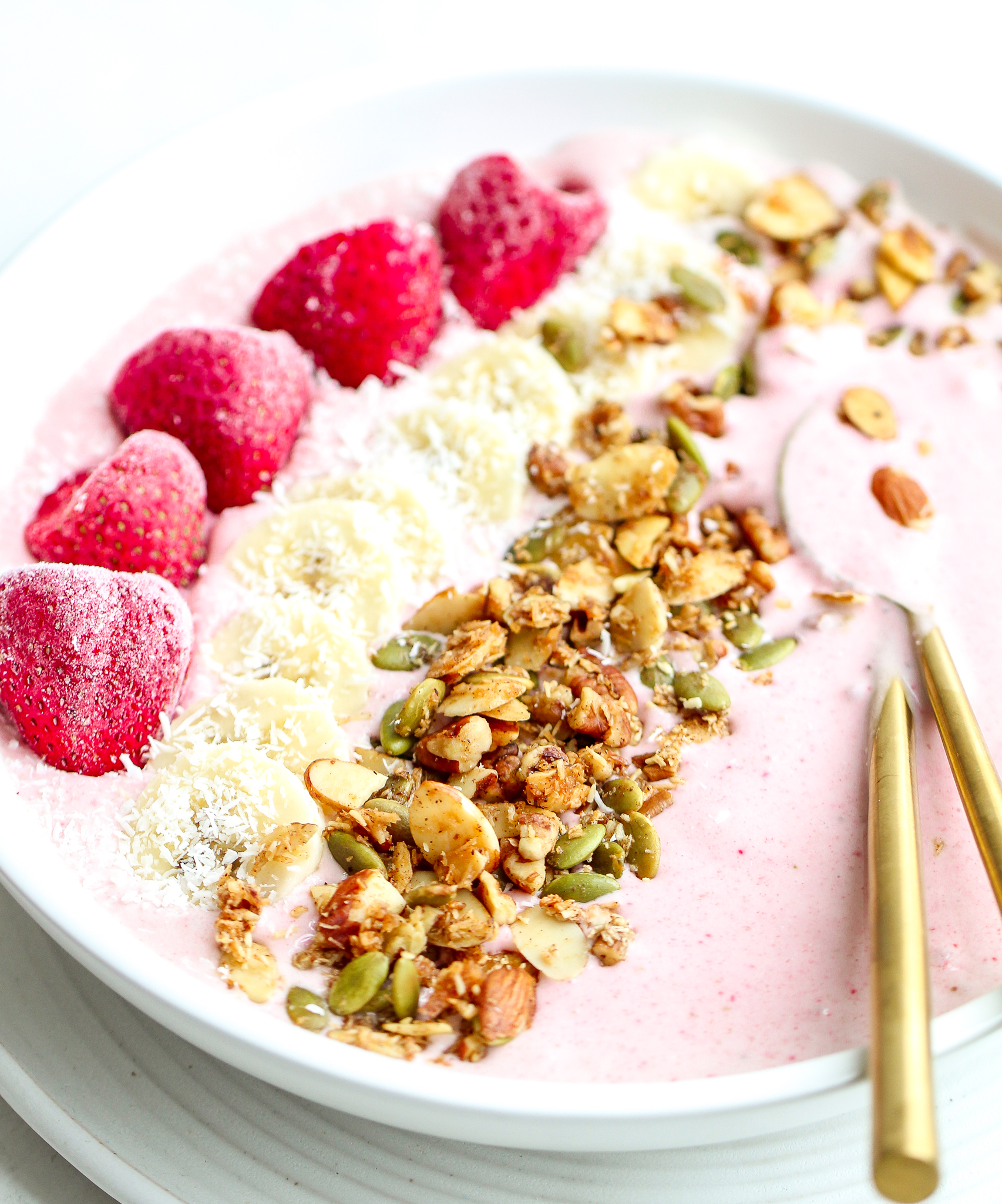 strawberry banana smoothie in a white bowl, topped with strawberries, sliced bananas, homemade granola and shredded coconut. There are matte golden spoons in the smoothie bowl 
