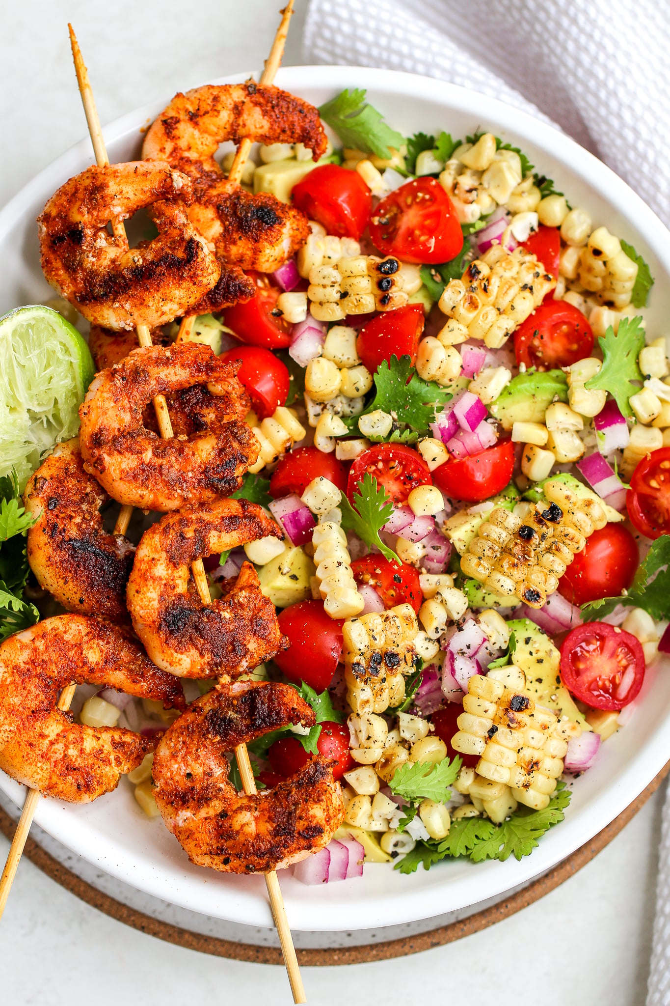 spiced grilled shrimp on wooden skewers laid over a white bowl layered with grilled corn, avocado, tomato, red onion and cilantro 
There is a squeezed lime on the side of the bowl 