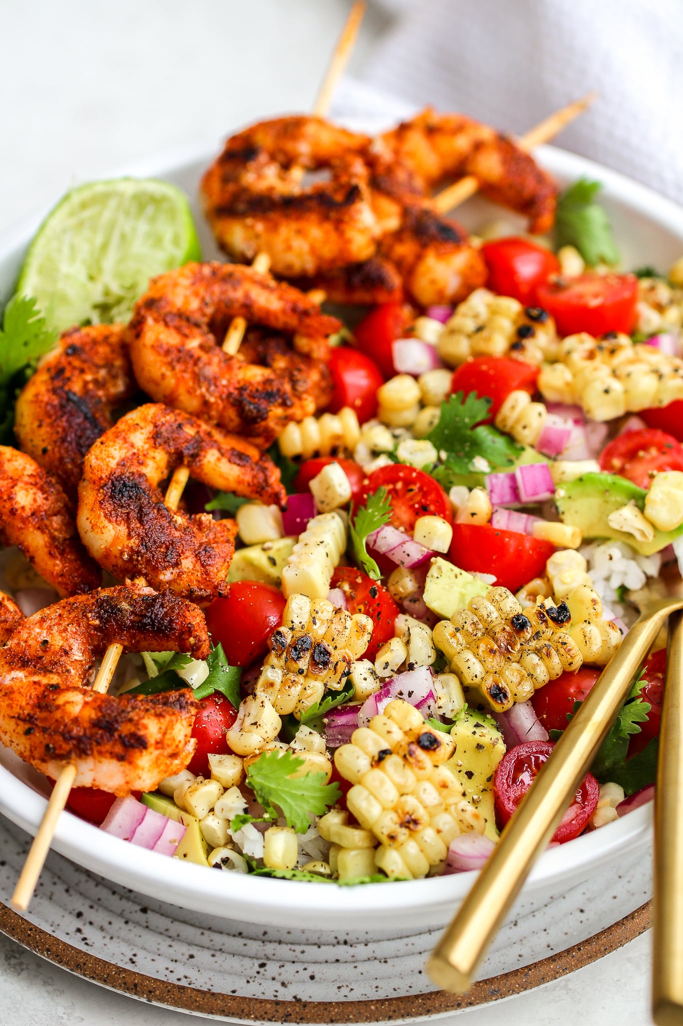 spiced grilled shrimp on wooden skewers laid over a bowl of fresh grilled corn and a variety of fresh veggies - diced red onion, cilantro leaves, grape tomatoes, avocado and lime