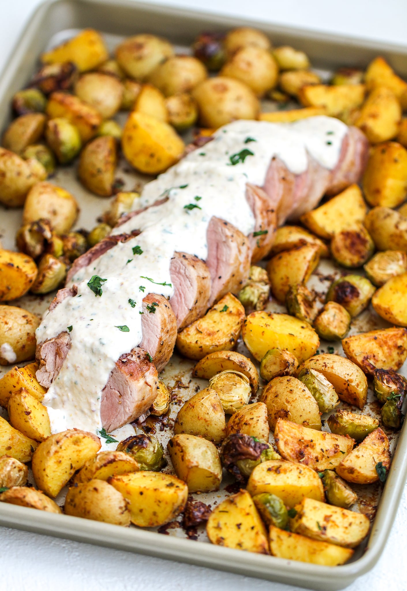 large sheet pan with cooked pork tenderloin in the center topped with creamy mustard sauce. there are crispy golden potatoes and Brussels sprouts surround the pork tenderloin