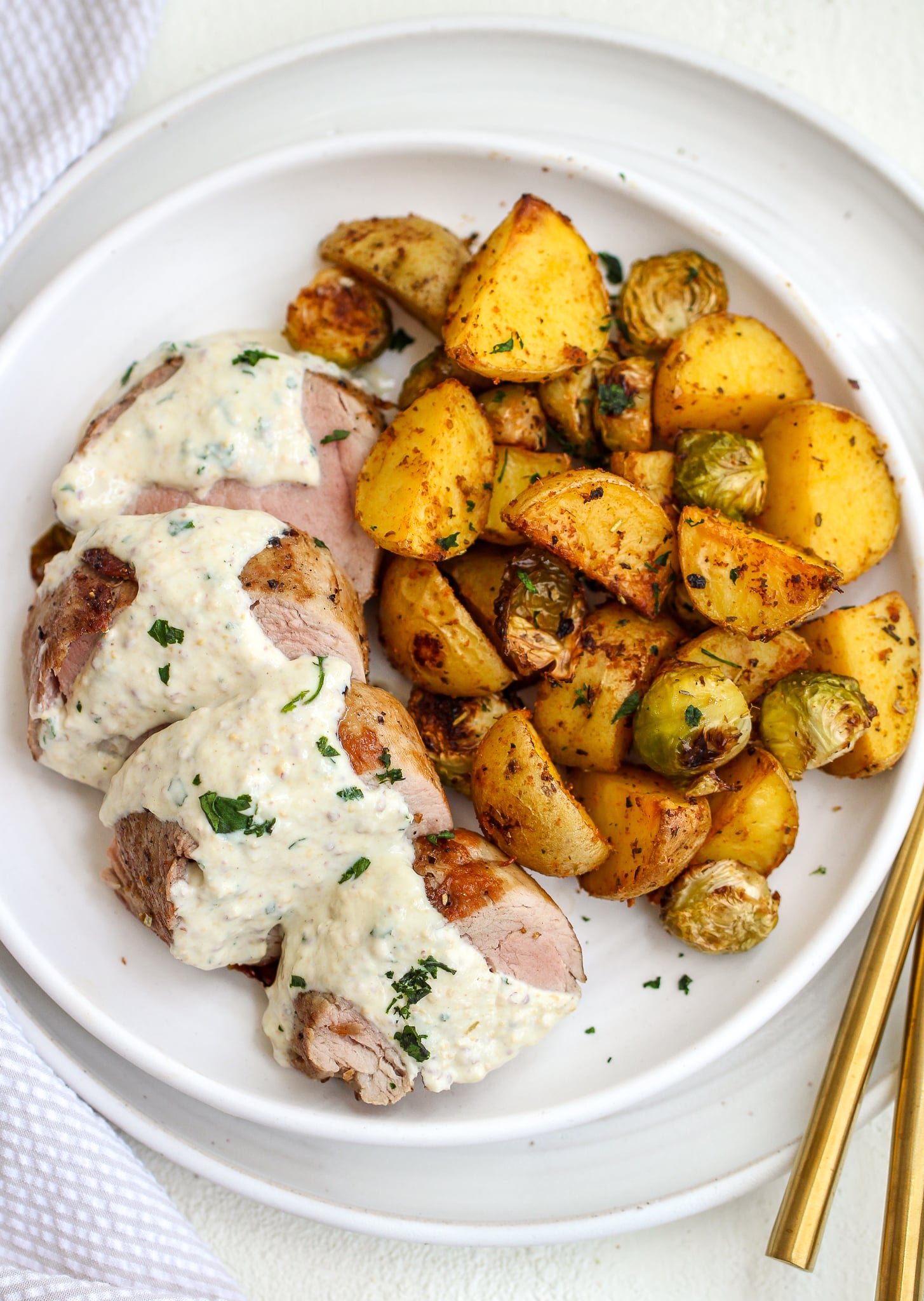cooked and slice pork tenderloin on a white ceramic dinner plate. the pork tenderloin is topped with creamy mustard sauce and there are crispy golden potatoes and Brussels sprouts on the side 