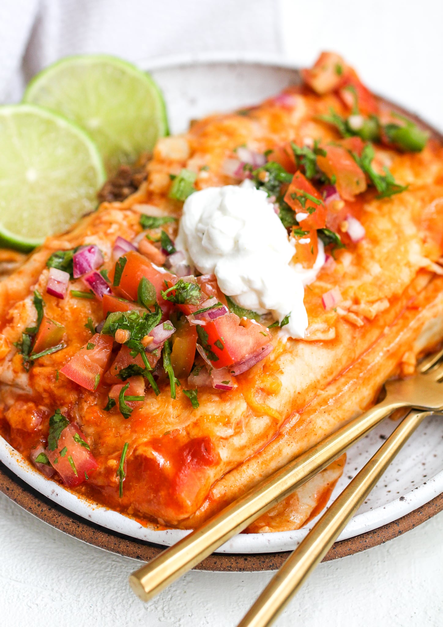 healthy enchiladas on a white speckled ceramic plate with pico de Gallo and sour cream on top. there are lime slices and gold forks on the side