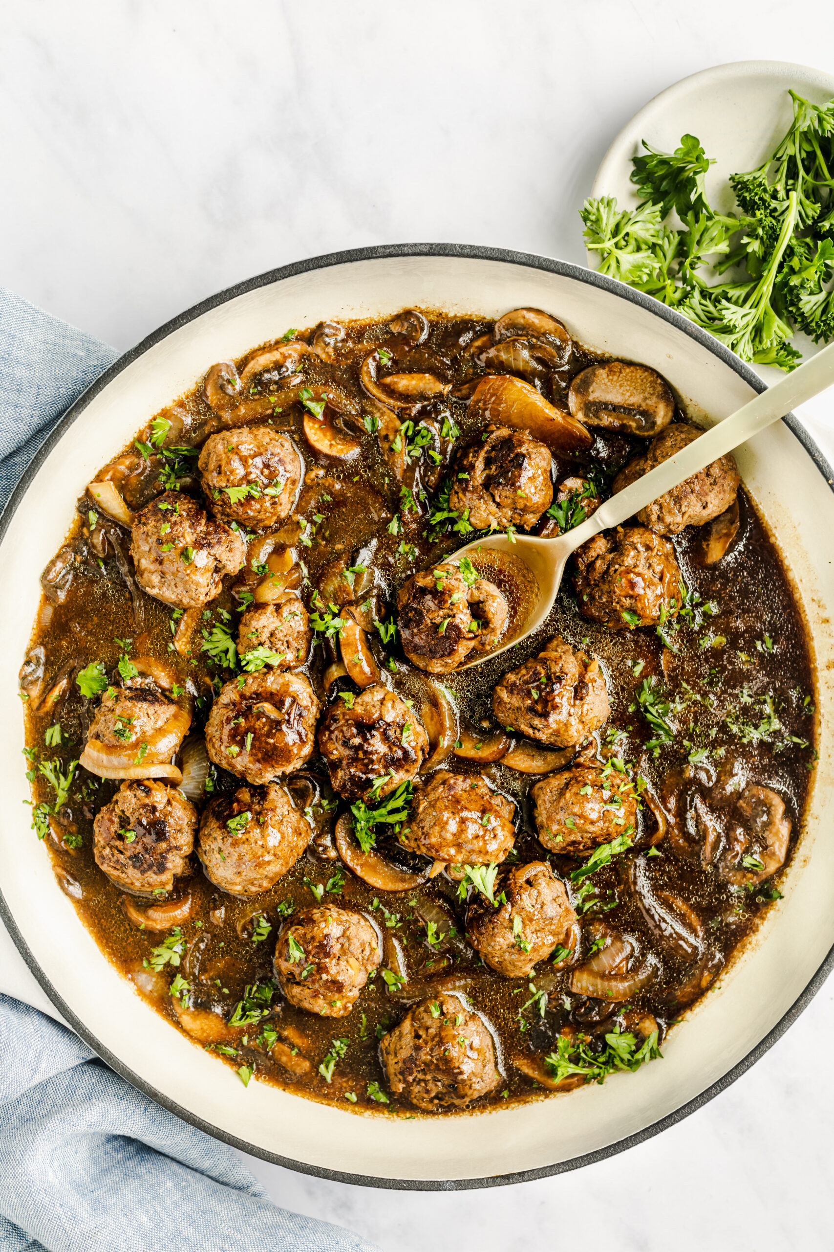 Salisbury steak meatballs in a large ceramic coated cast iron skillet with a serving spoon in it - topped with fresh parsley