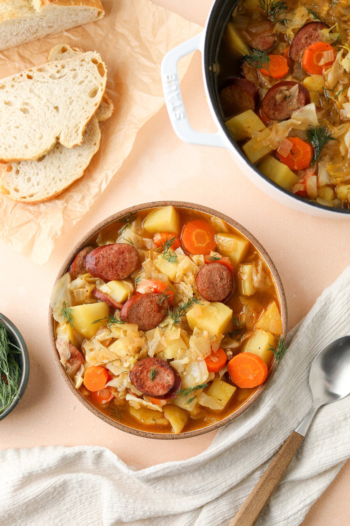 Overhead photo of kielbasa and cabbage soup in a serving bowl - it is served alongside sliced bread and fresh dill