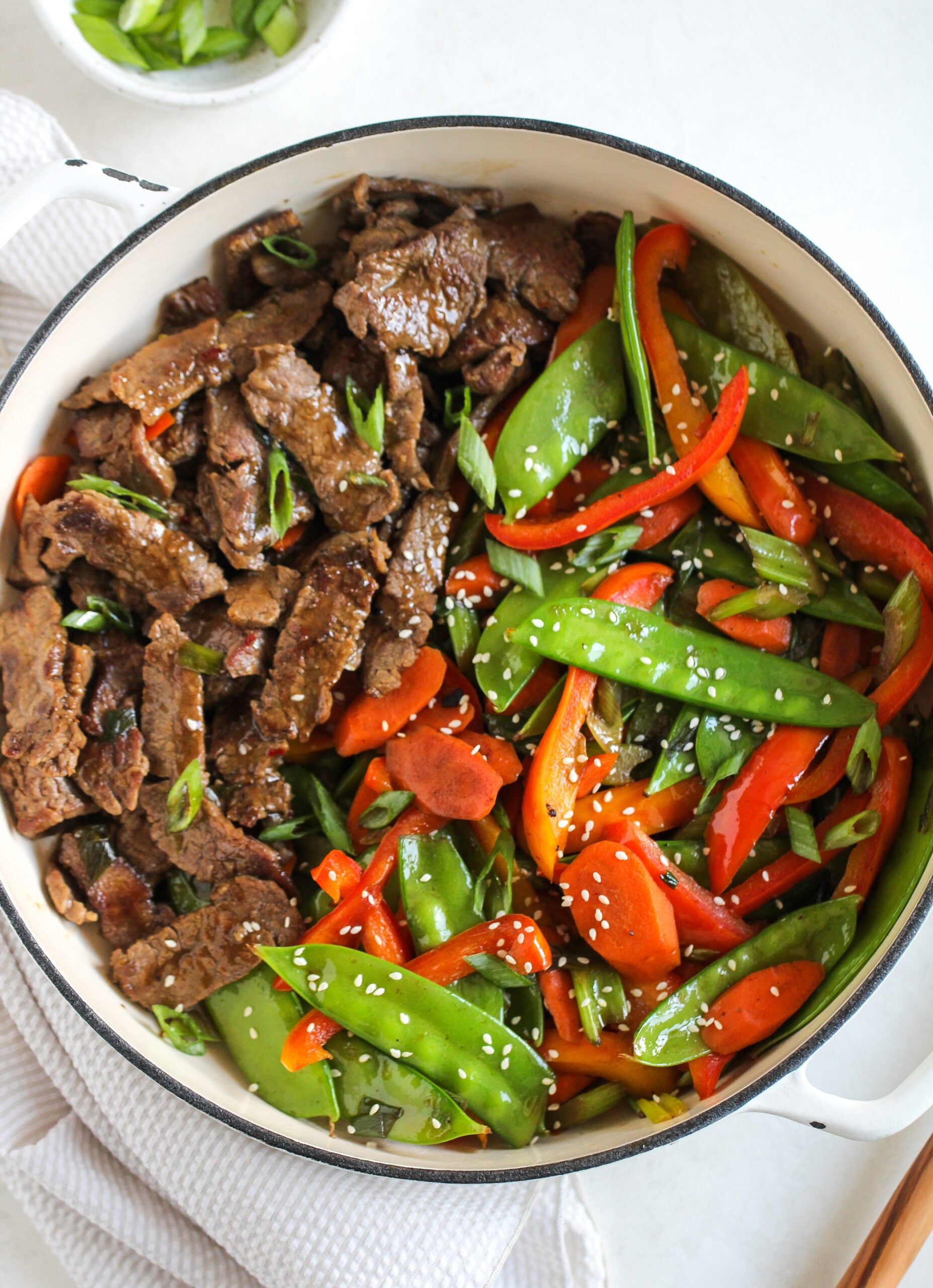 beef and veggie stir fry in a large white skillet - topped with white sesame seeds and fresh green onions cut on the diagonal 