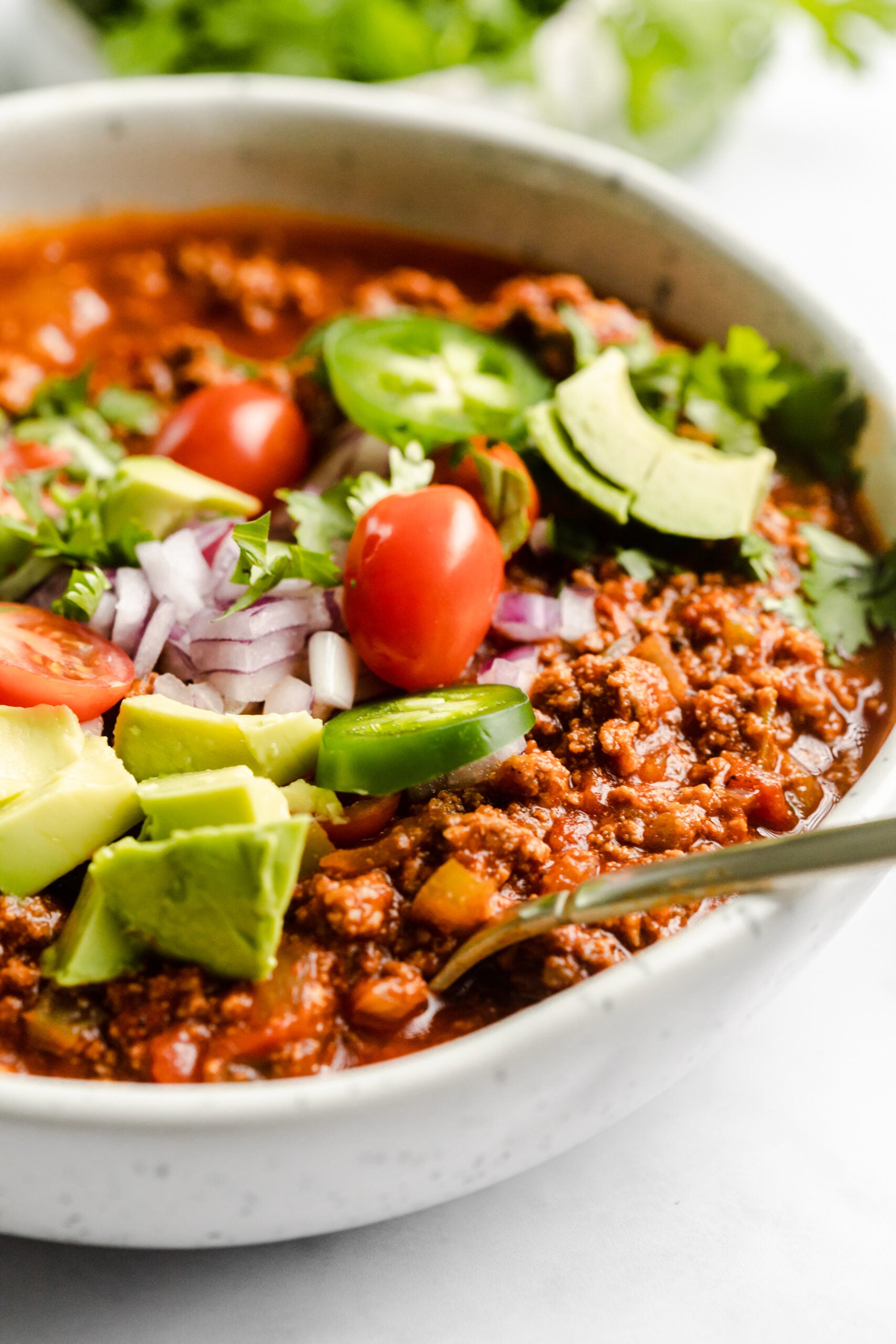 close up photo of no bean chili in a bowl topped with fresh tomatoes, diced red onion, sliced jalapeños and cilantro 
There is a spoon sticking out of the bowl 