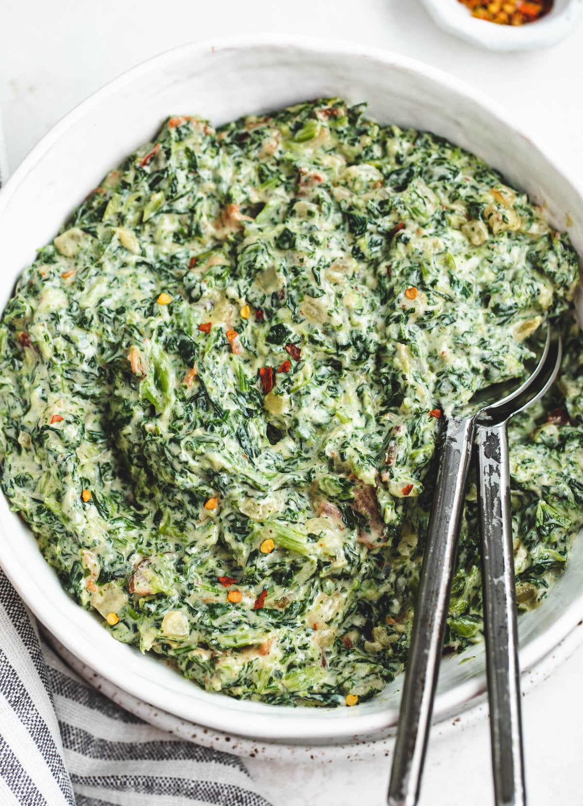 White serving bowl with creamed spinach. It has two serving spoons in it and is topped with a few crushed red pepper flakes. 