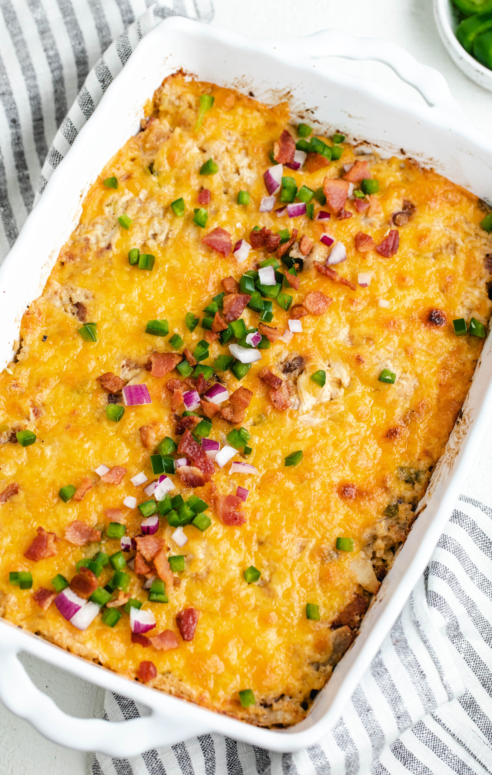 Cheesy jalapeño popper casserole in a white casserole dish, it is topped with diced fresh jalapeño, red onion and bacon bits.
