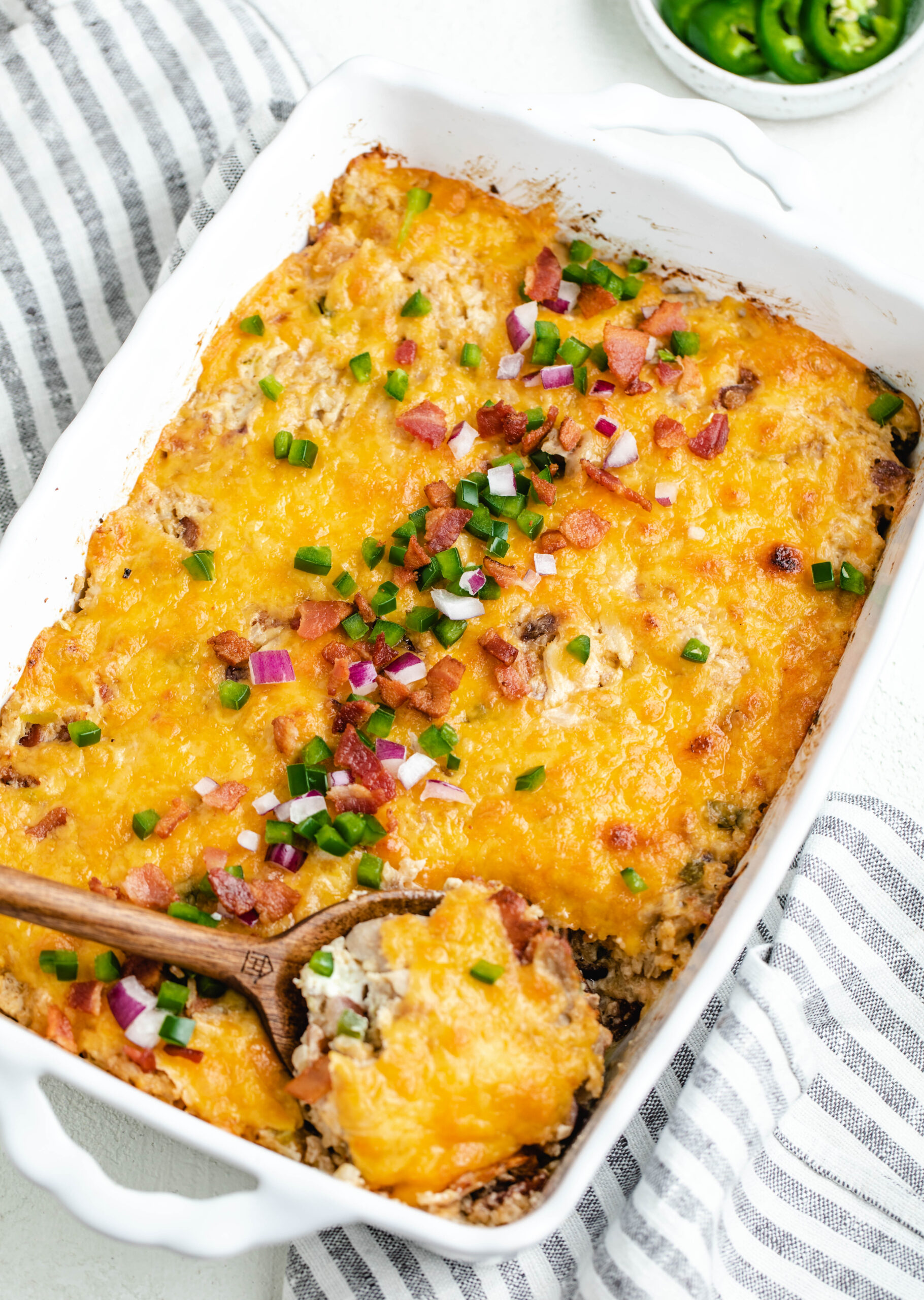 serving spoon taking a scoop out of the jalapeño popper casserole. the casserole is topped with diced jalapeños, red onion and chopped bacon.