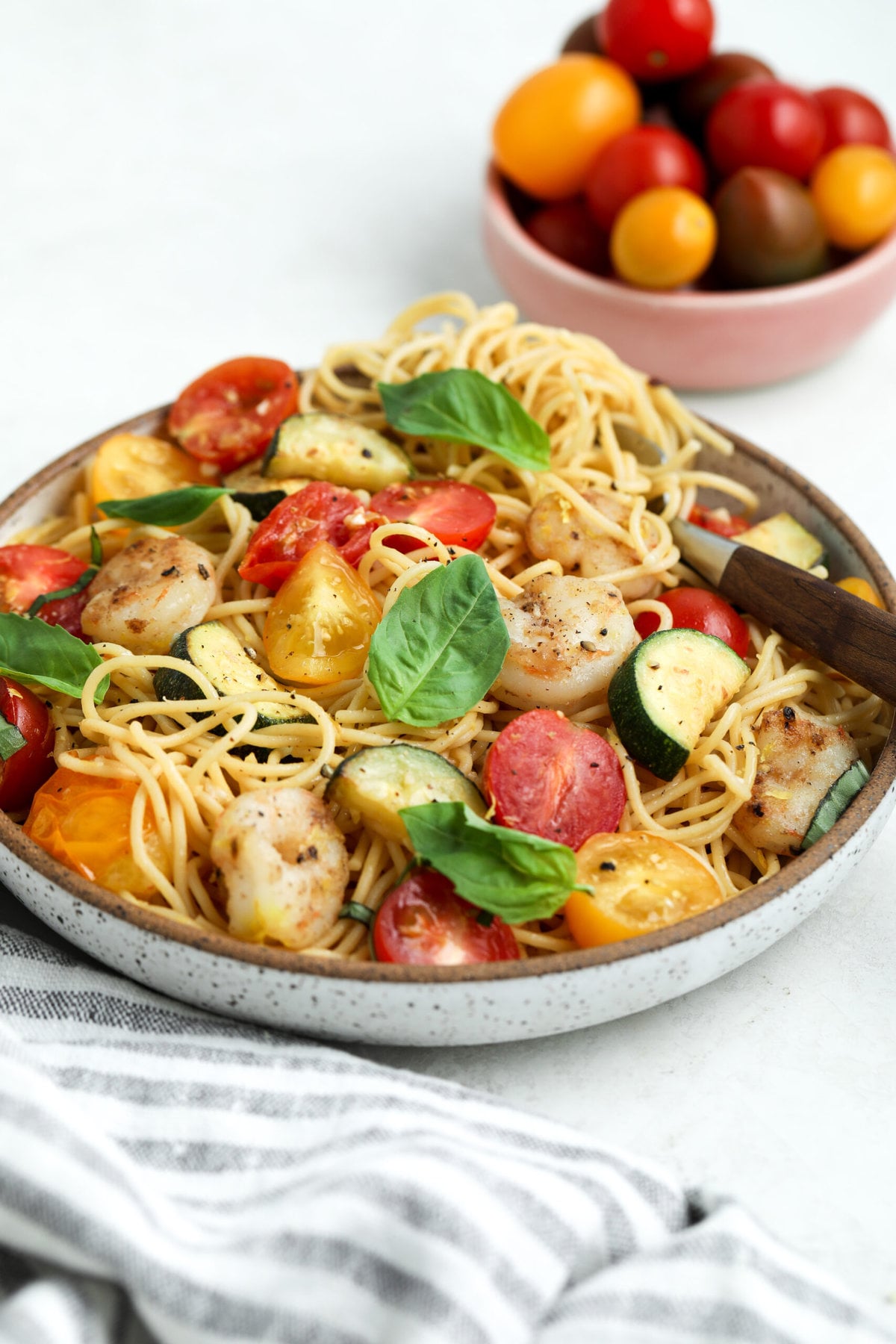 tomato and zucchini pasta in a serving bowl. there is a fork twirled into the noodles and the pasta is topped with fresh basil and cracked pepper