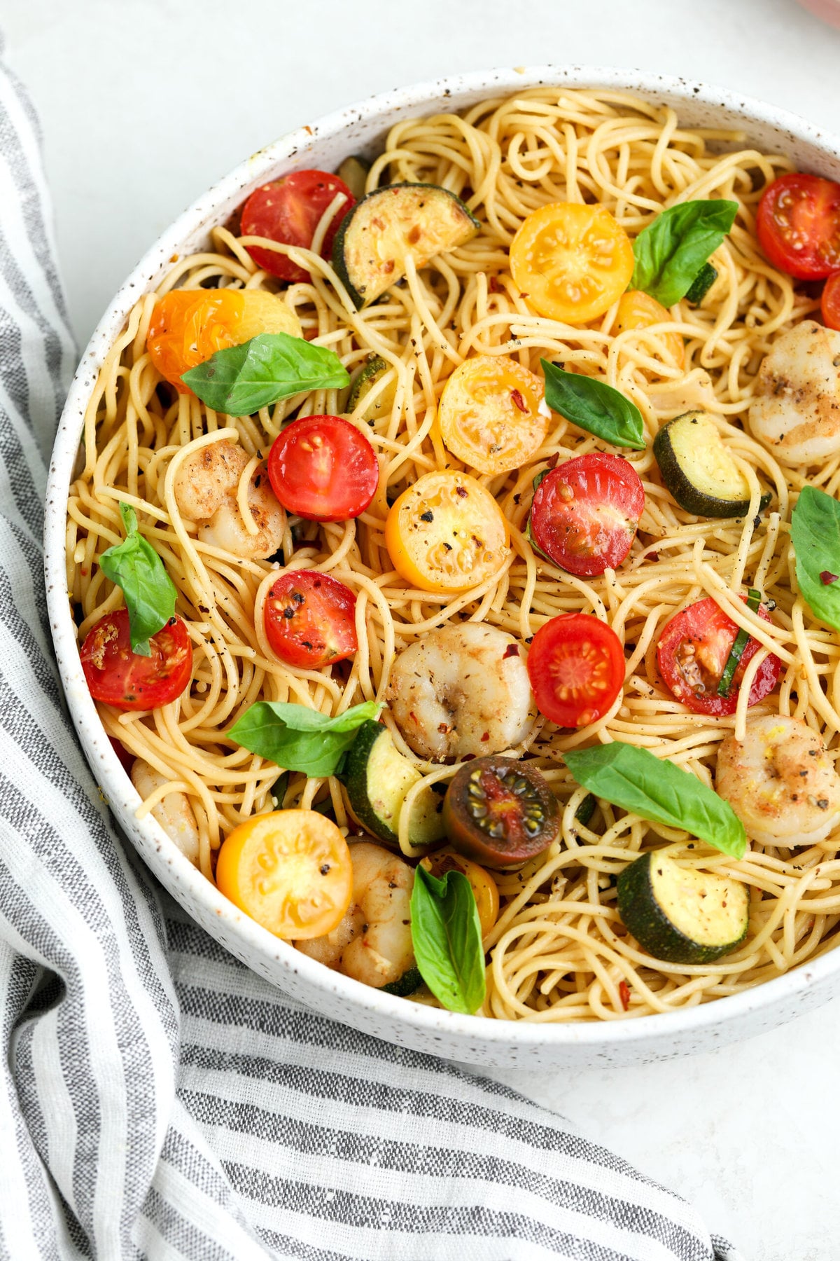 large ceramic serving bowl filled with tomato and zucchini pasta. It is topped with fresh basil and cracked pepper. There is a blue and white striped kitchen towel next to it.