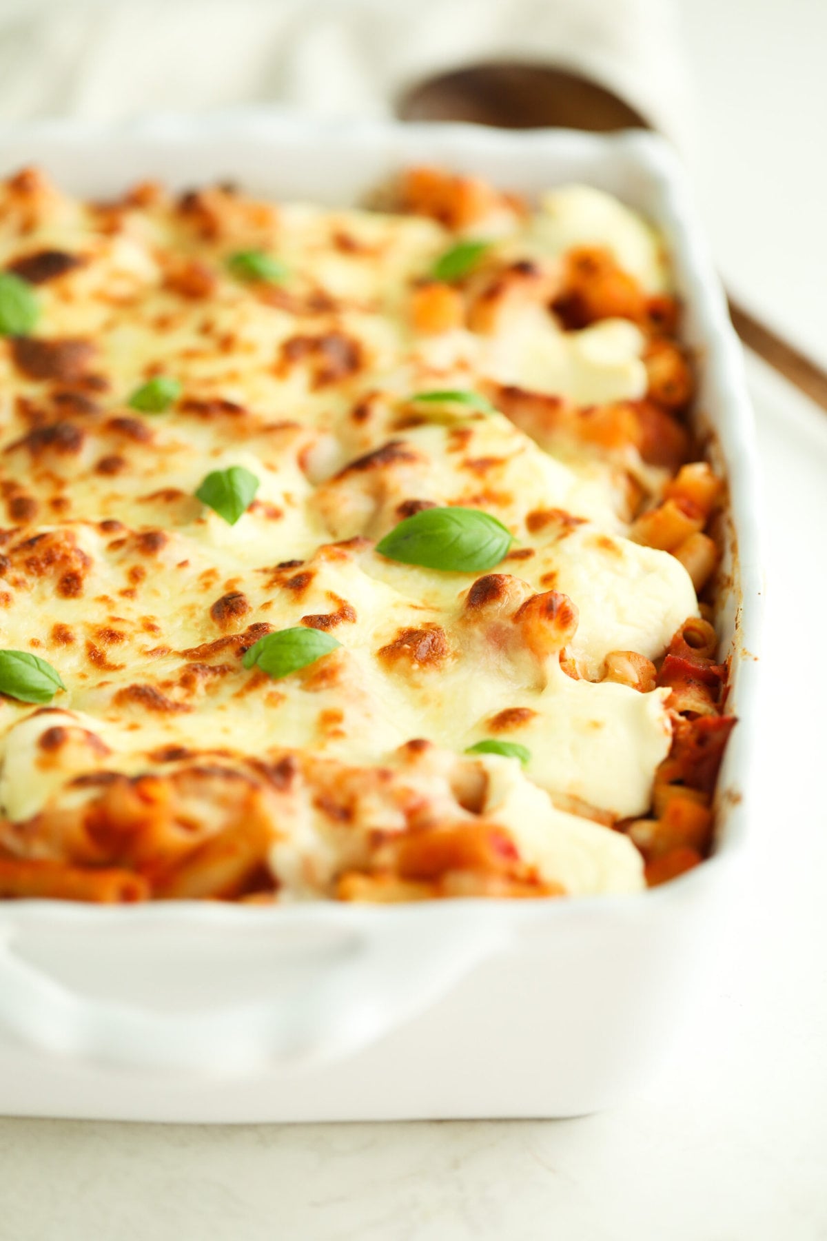 side view of baked ziti in a white baking dish - it is topped with melty cheese and fresh basil leaves